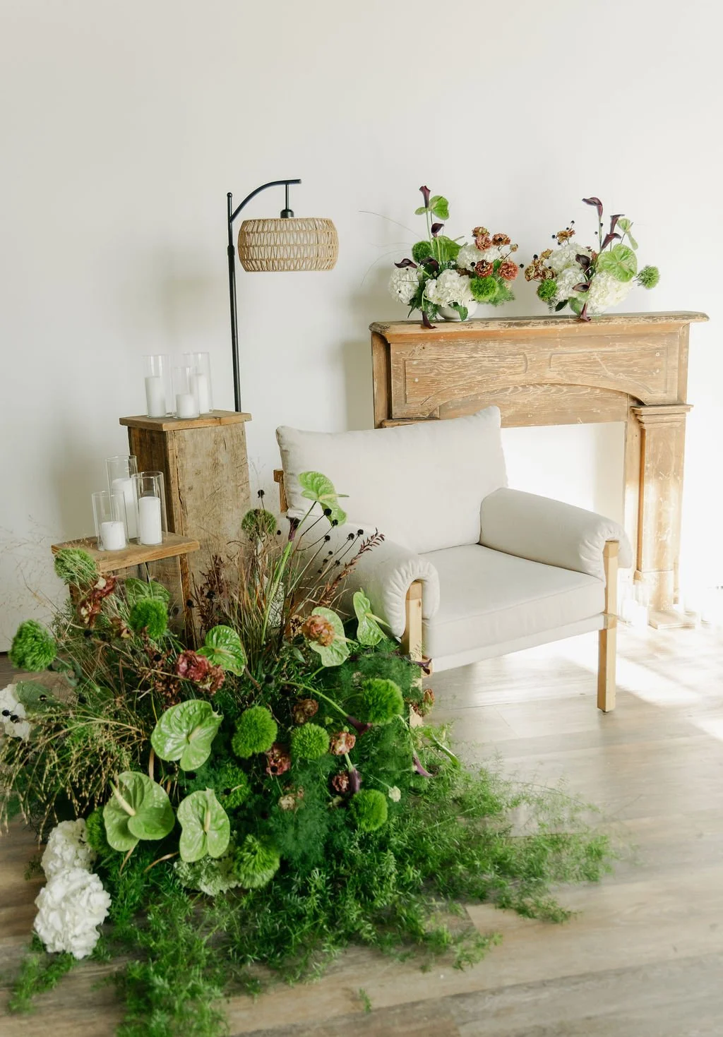 Light-filled living room with a white armchair, wooden fireplace mantel decorated with flowers, large greenery arrangement on the floor, wooden side tables with candles, and a black floor lamp with a woven rattan shade.