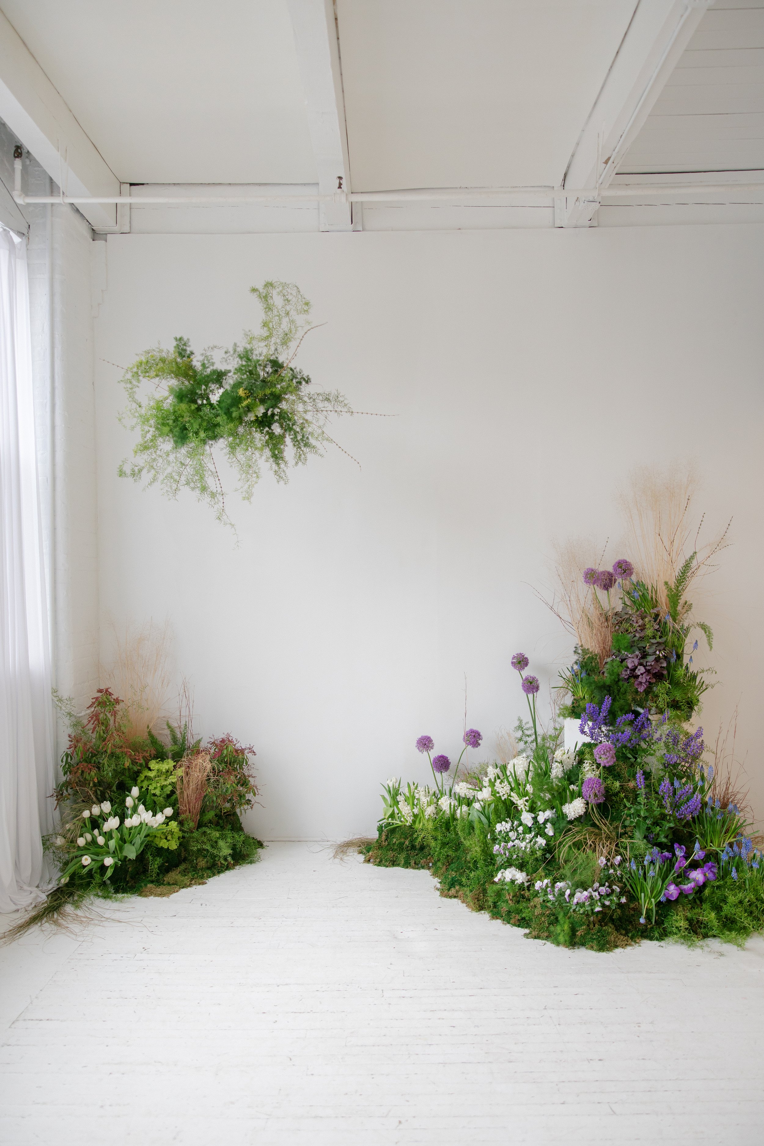 A white indoor space decorated with a variety of colorful flowers and green plants arranged along the bottom and side edges, and a hanging greenery installation in the upper center against a white wall and ceiling at The George Eastman House.