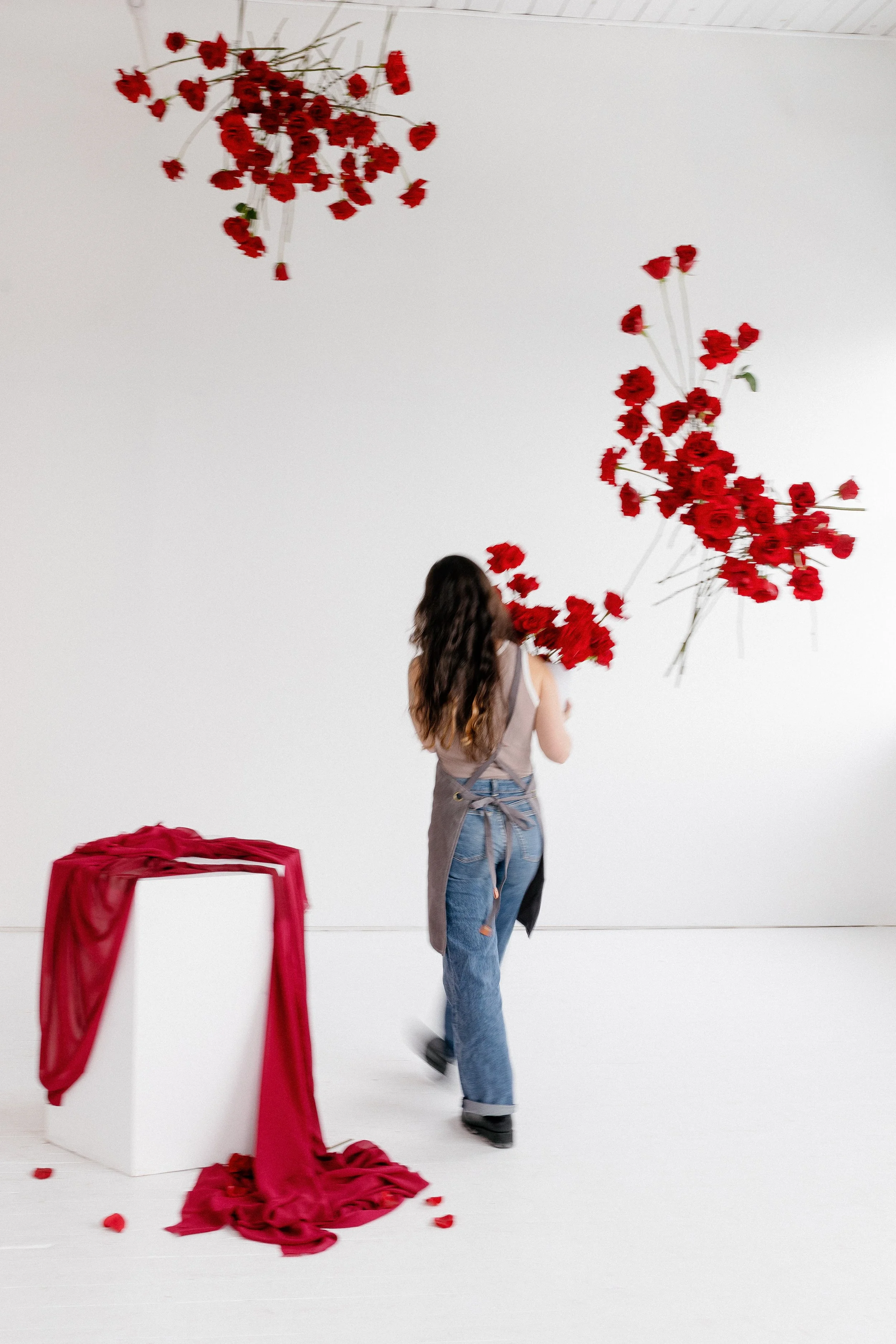 A woman with long brown hair, dressed casually in a gray top and jeans, stands with her back to the camera in front of a white wall. Red roses are suspended on strings from the ceiling to the right and above her. A white pedestal with a red cloth dra