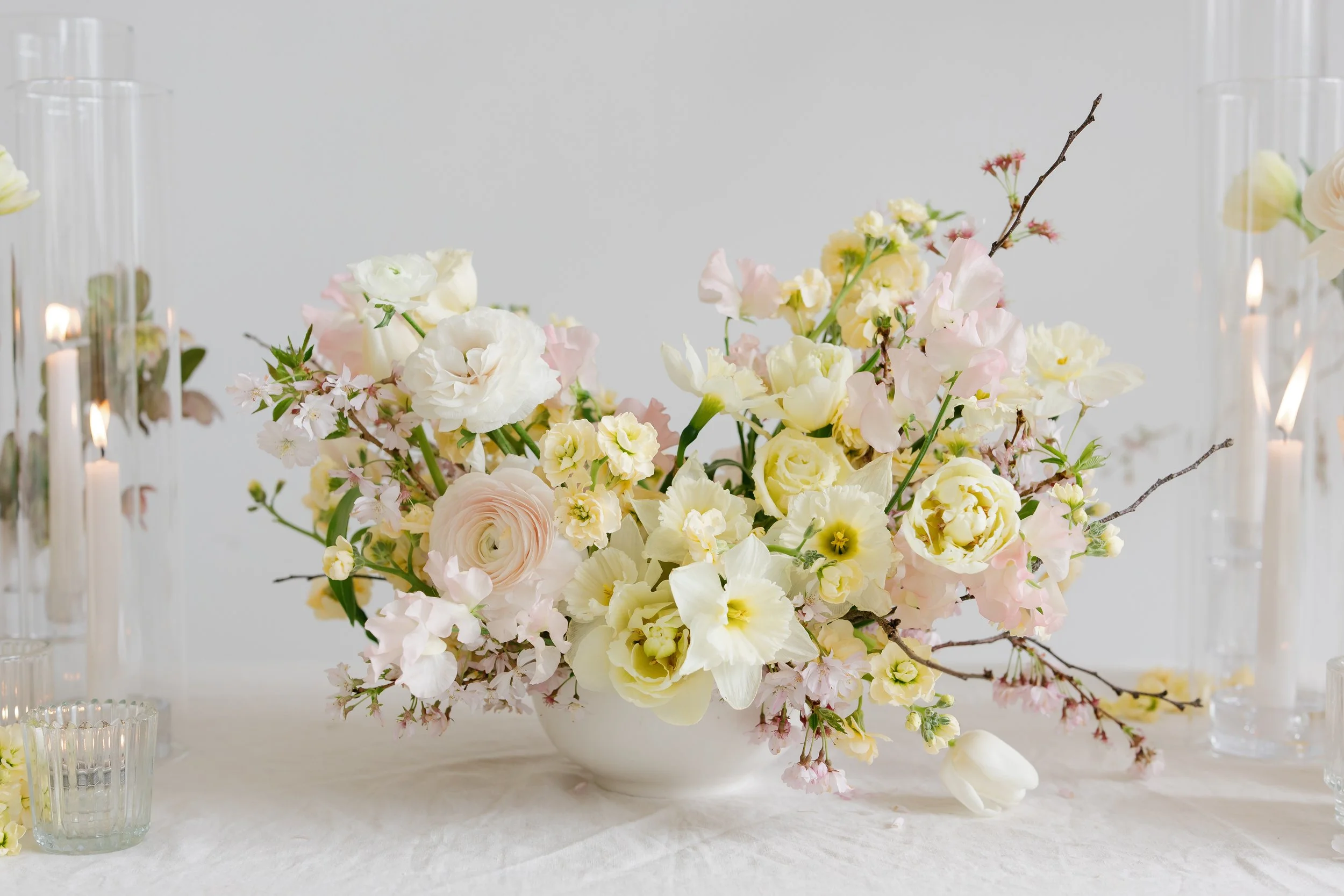 Elegant floral centerpiece with white, cream, and light pink flowers on a table with glass candle holders and a white background setting up in Buffalo, NY  for a corporate event.
