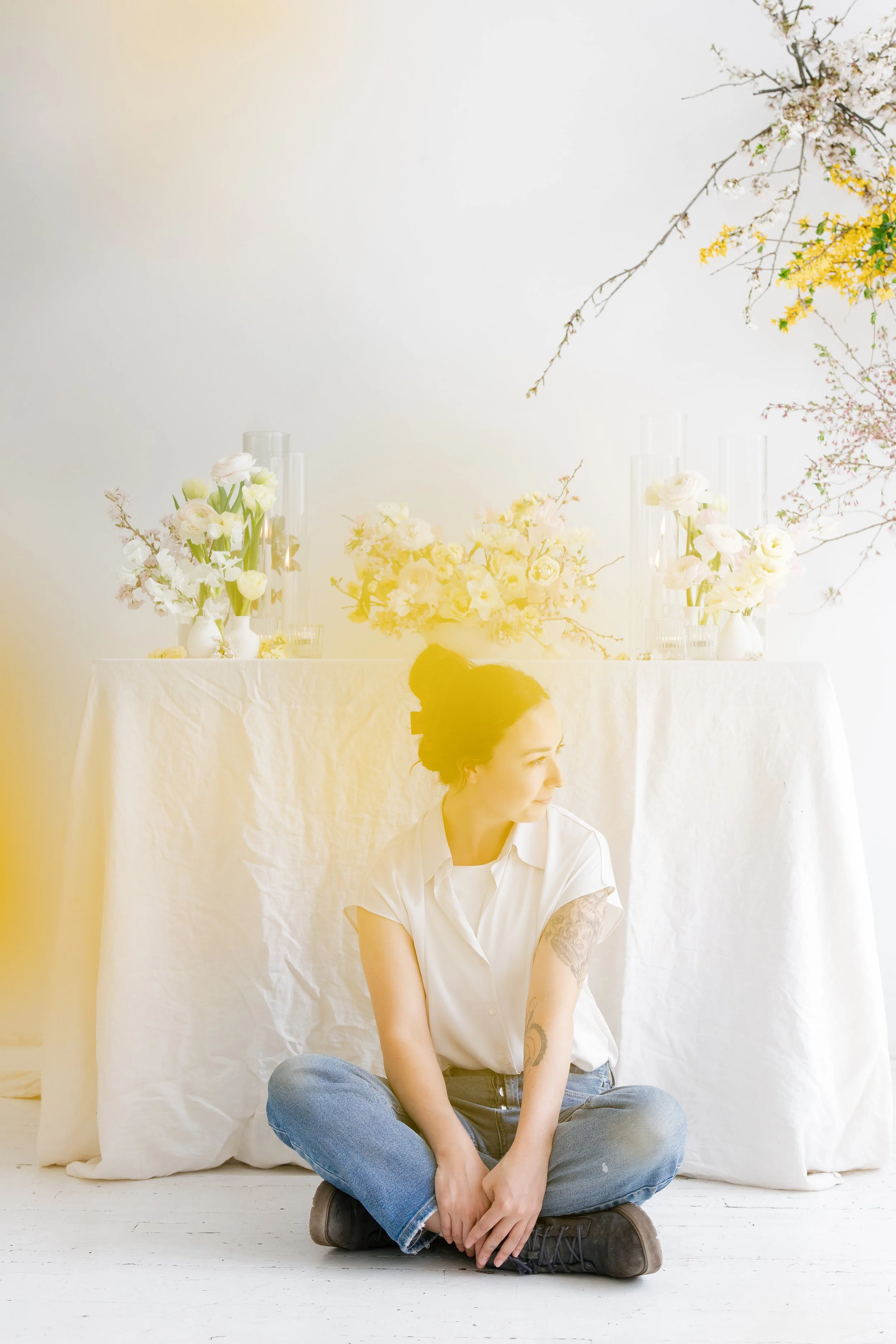 A woman with dark hair in a bun, wearing a white shirt and jeans, sitting cross-legged on the floor in front of a white table with yellow and white floral arrangements on it, against a white background with branches of flowers on the top right.