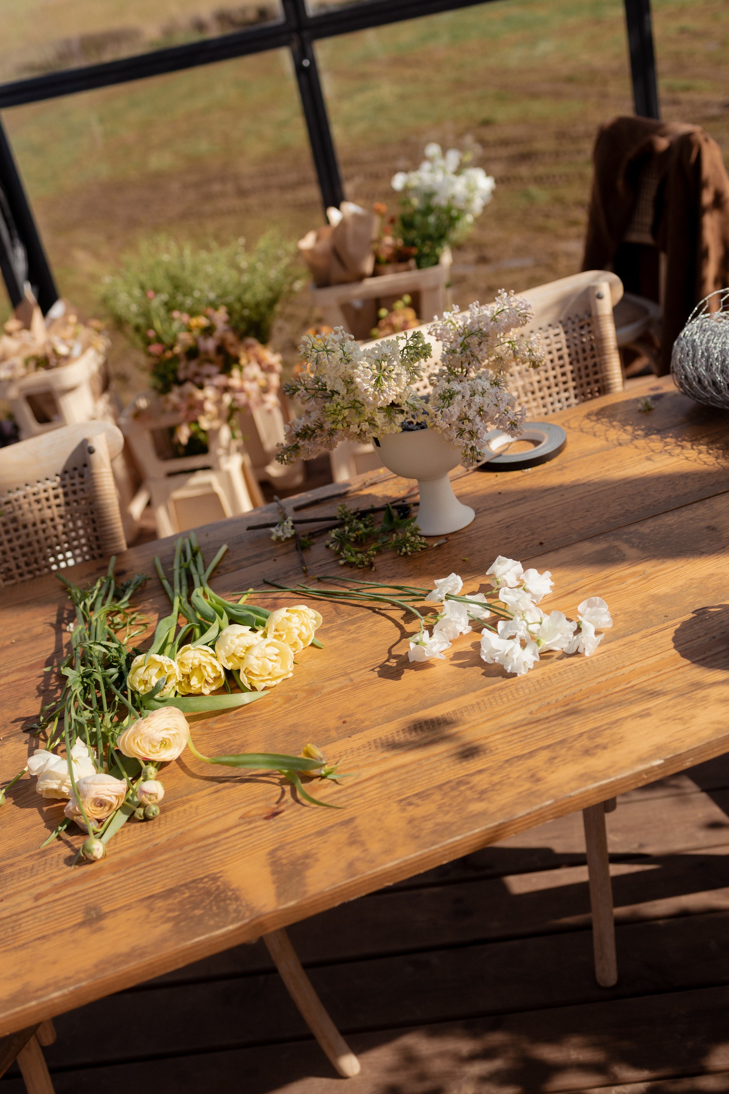 A wooden table decorated with white and yellow flowers, set in a bright outdoor space with chairs and potted plants at The Orangery with Trille Floral in Bath, United Kingdom.