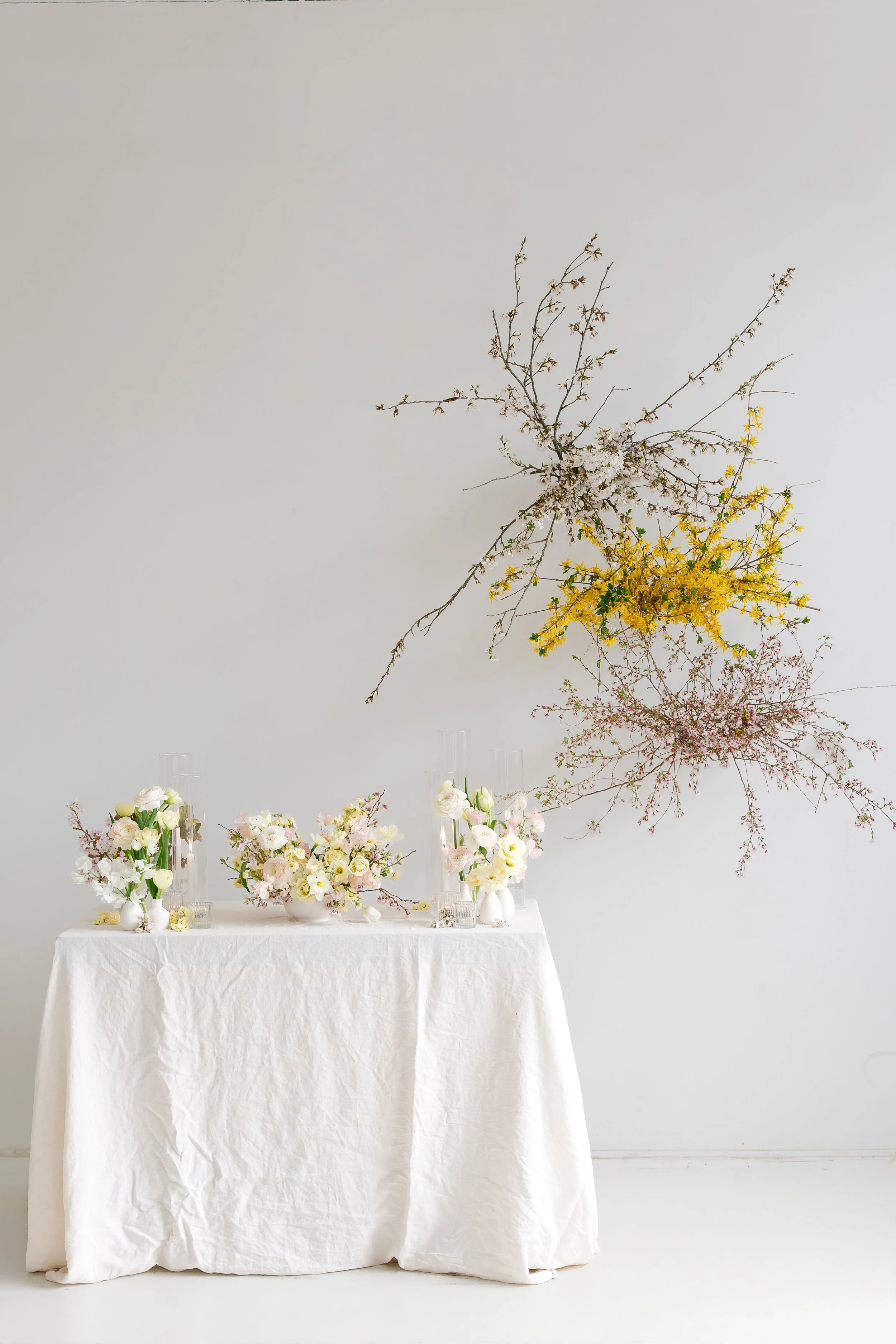A table covered with a white tablecloth displaying multiple floral arrangements in white vases and bowls, with creamy and pastel-colored flowers and tall, clear glass vases, against a plain light grey wall decorated with large, branching floral arran