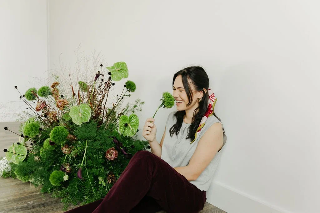 Woman sitting on the floor next to a large arrangement of green and purple flowers, holding a green flower close to her face, smiling.