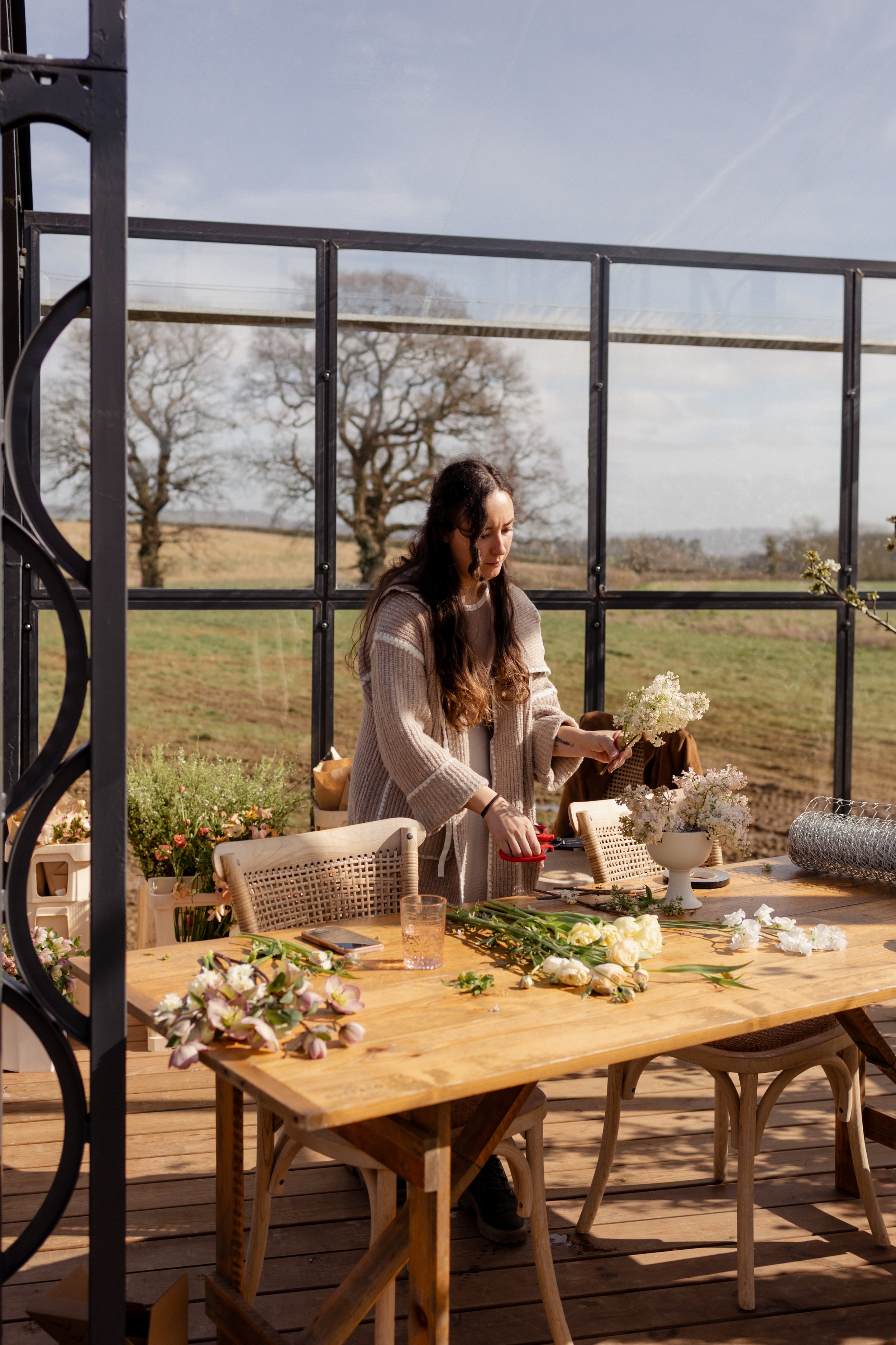 A woman arranging flowers at a table outdoors on a sunny day, with a rural landscape of trees and fields in the background for Trille's Floral Masterclass.