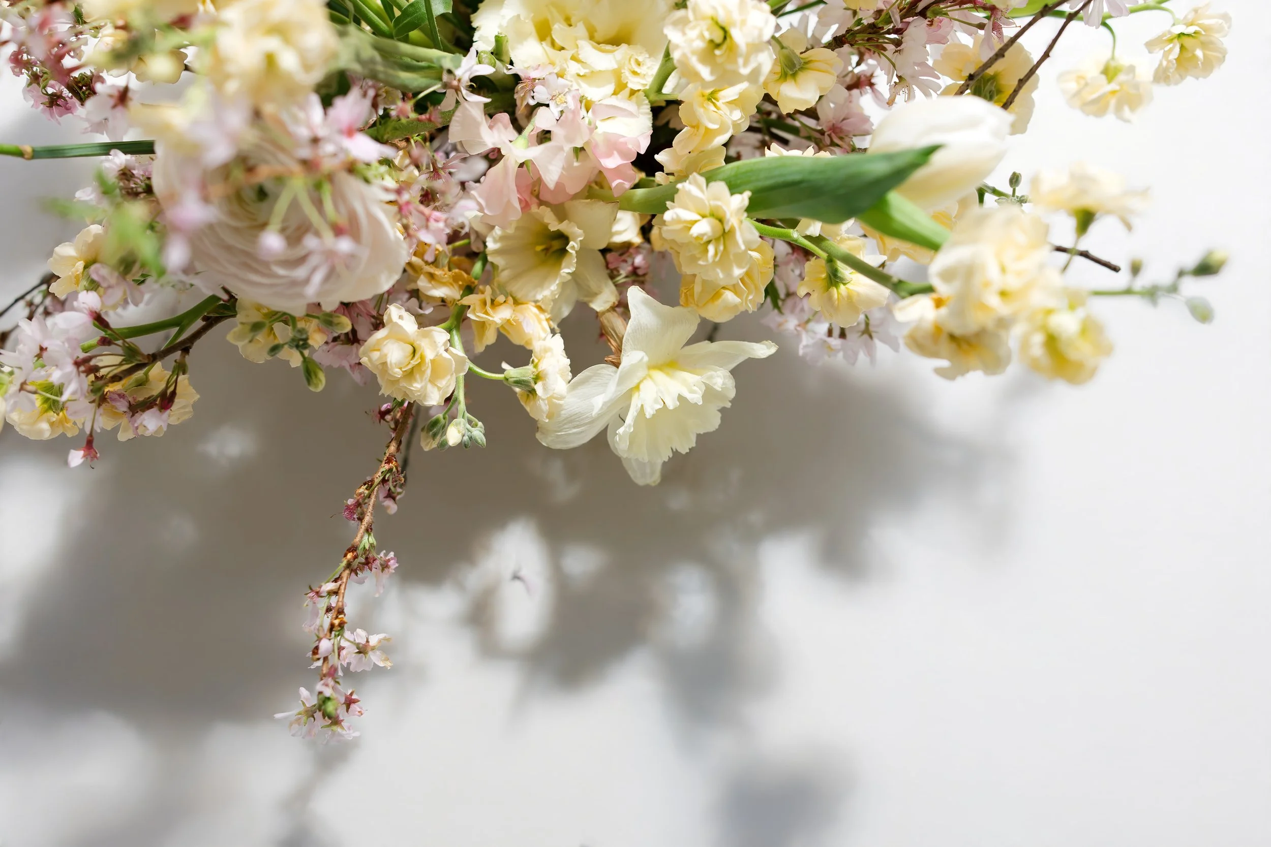 Close-up of a bouquet of white, cream, and light pink flowers with green leaves against a light background.