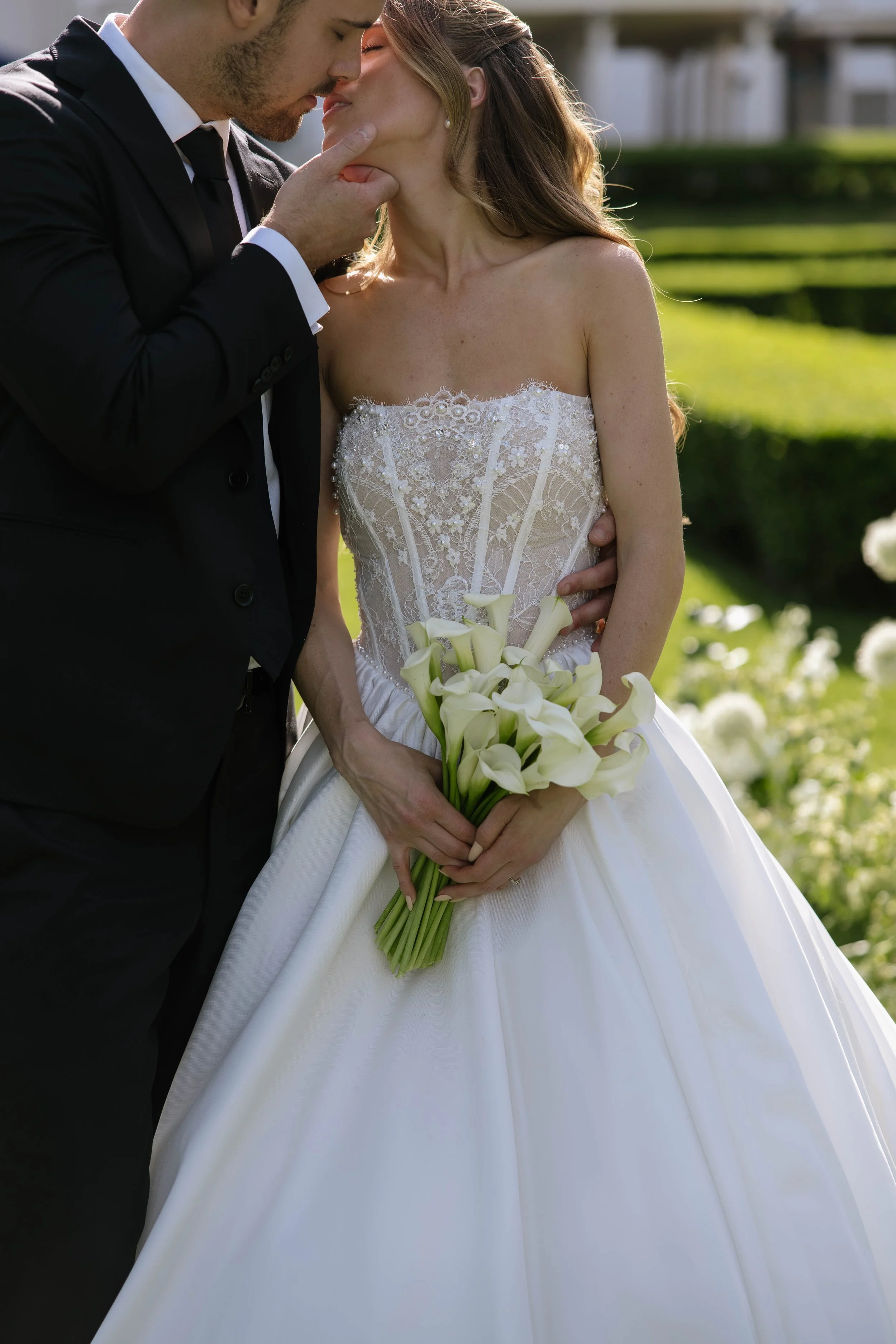 A bride and groom share a close moment outdoors. The bride holds a bouquet of white calla lilies, wearing a white lace wedding dress, and the groom wears a black tuxedo. They are touching foreheads with their eyes at Geneva on the Lake