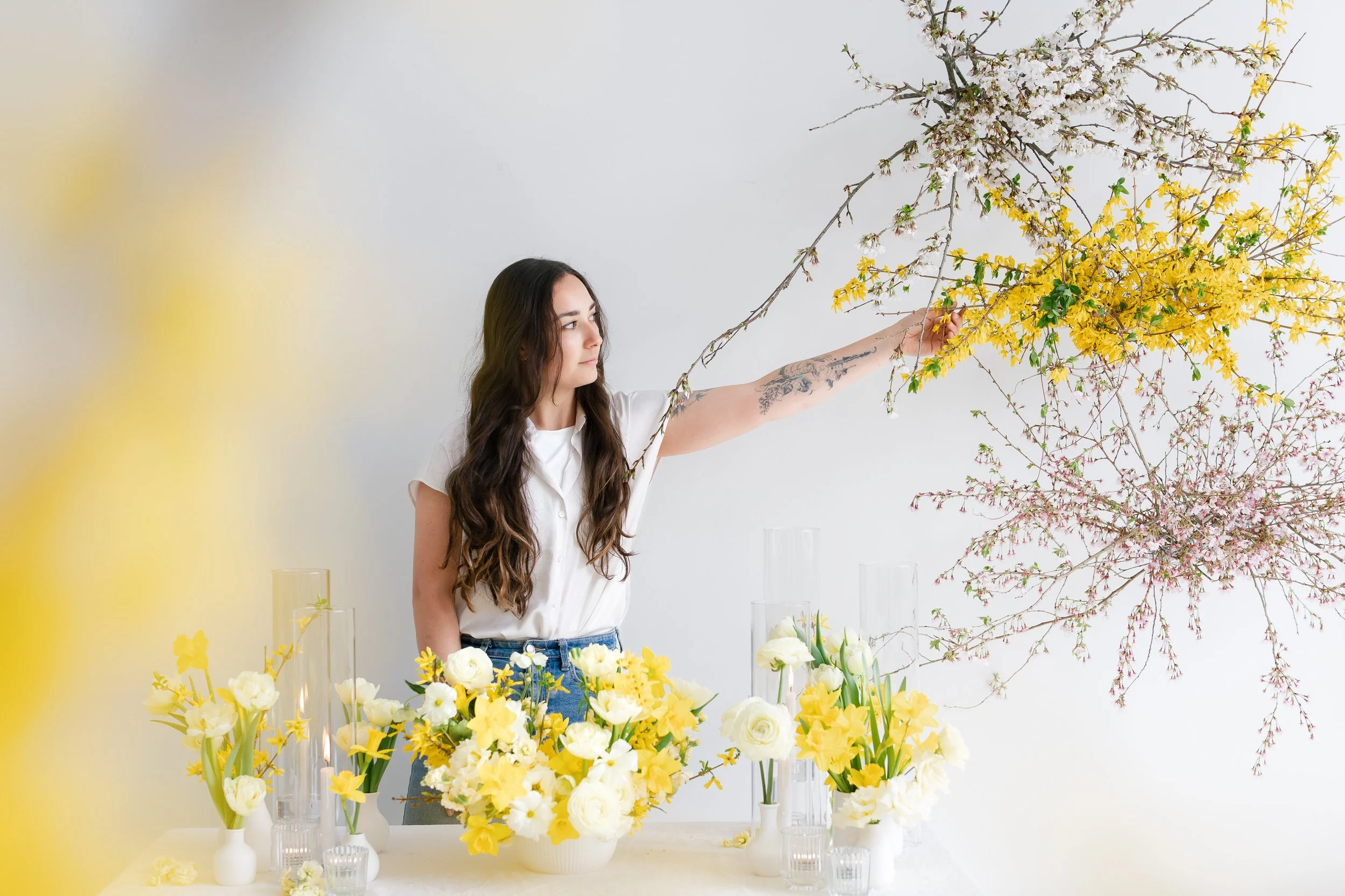 Midnight Moss arranging yellow and white flowers in a hanging installation, with cherry blossom and daffodil branches hanging above, against a plain white wall for a wedding ceremony in The Finger Lakes.