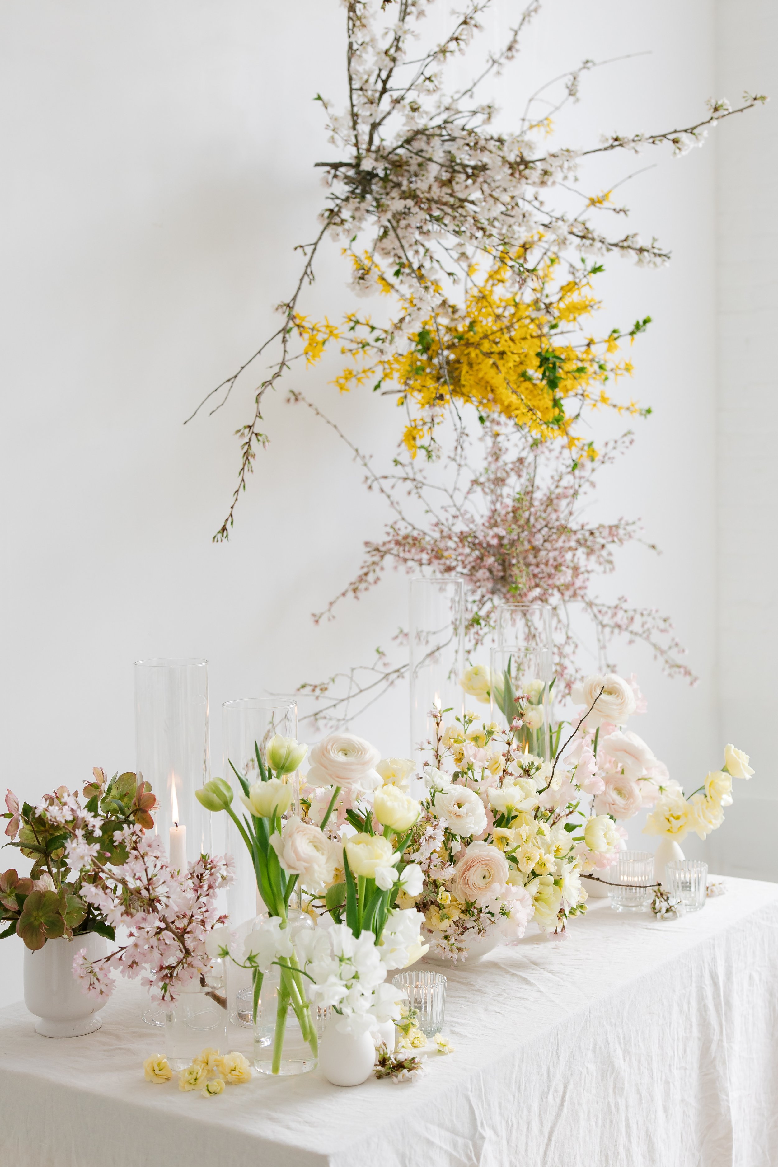 Luxurious floral arrangement with white daffodil, pink cherry blossoms, and yellow forsythia on a table with glass vases and candles, with a white background at Harro East Ballroom in Rochester, NY.