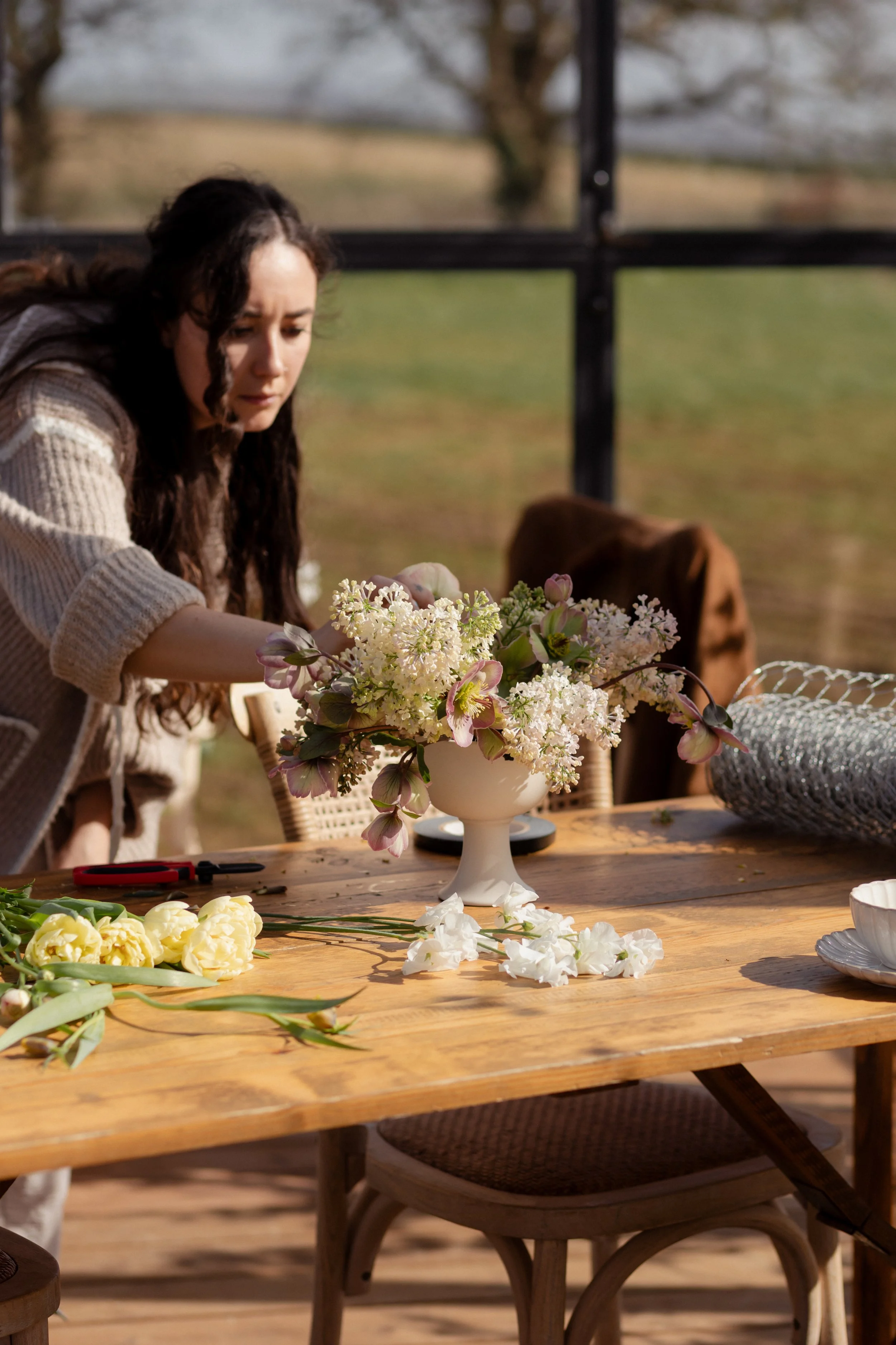 A woman with long dark hair arranging flowers on a wooden table outdoors during daytime. The table has various flowers, scissors, and floral arranging tools, and a large vase of mixed pink and white flowers is in the center. The background shows a gl