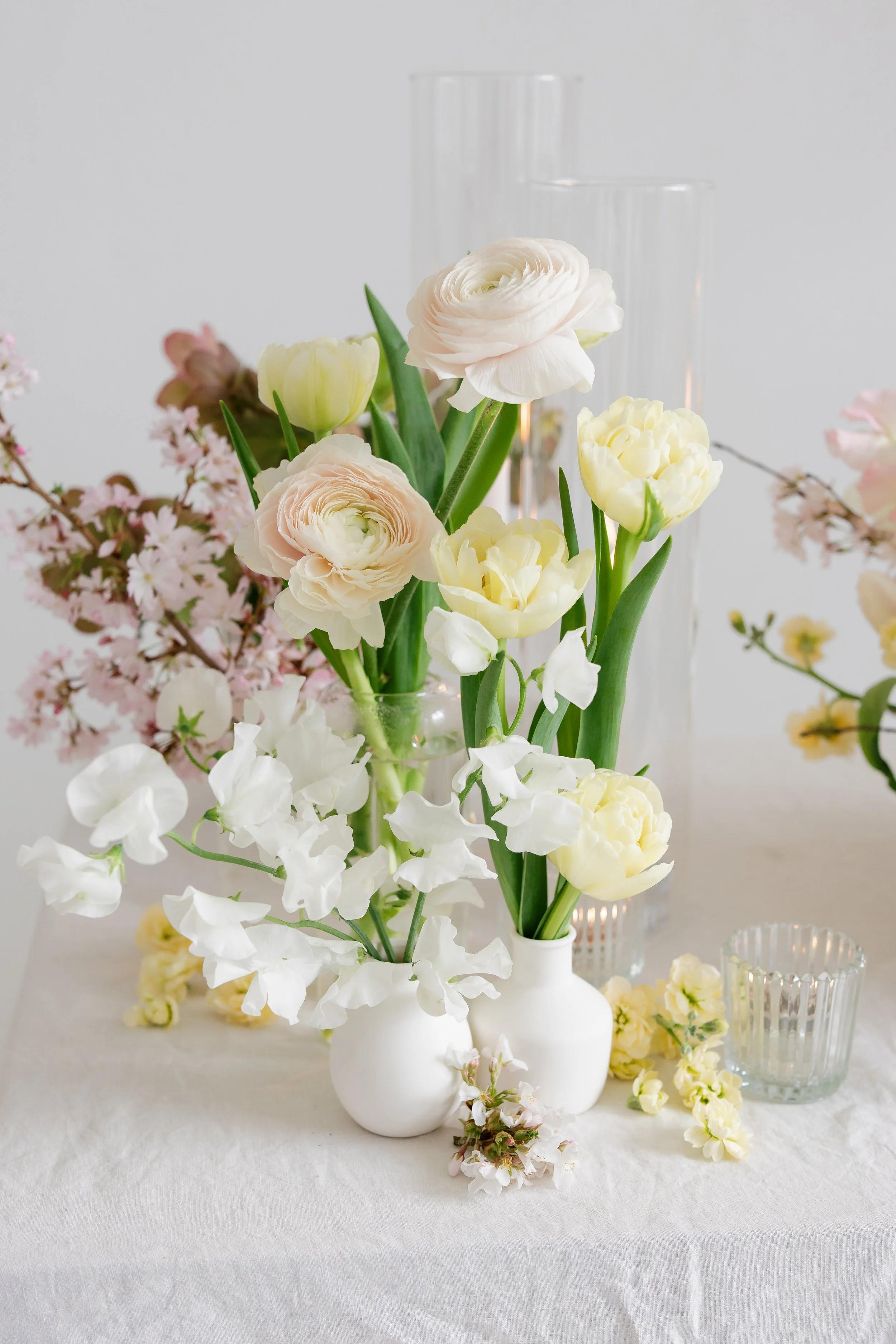 Elegant floral arrangement of white and cream flowers in white vases on a table with a light-colored tablecloth, complemented by glassware and small decorative items in the background shot by Jacalyn Meyvis Photography.