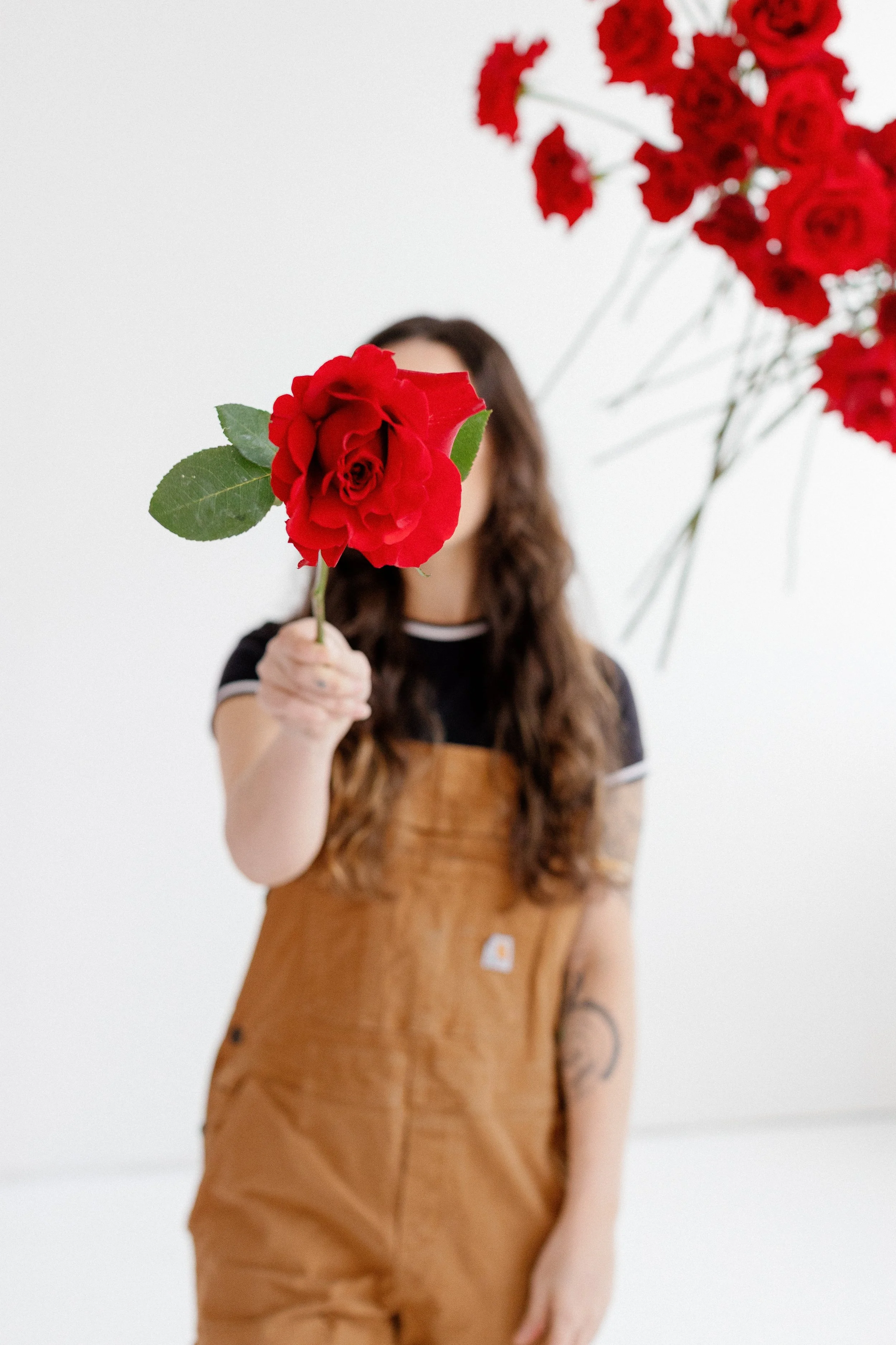 Person with long, wavy hair holding a red rose in front of their face, with blurred red flowers and a white background.