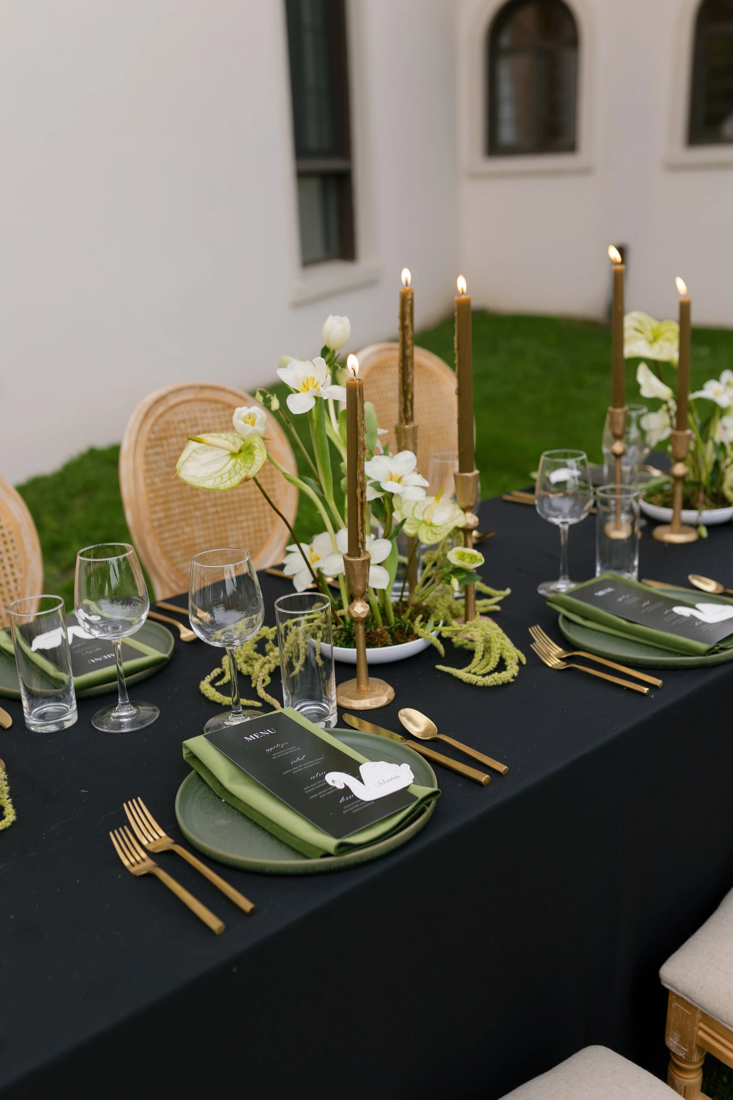 A formal outdoor dining table with black tablecloth, gold utensils, green napkins, clear glassware, ikebanas with white flowers and greenery, tall brown candles, menu cards, and beige woven chairs, at Geneva on the Lake