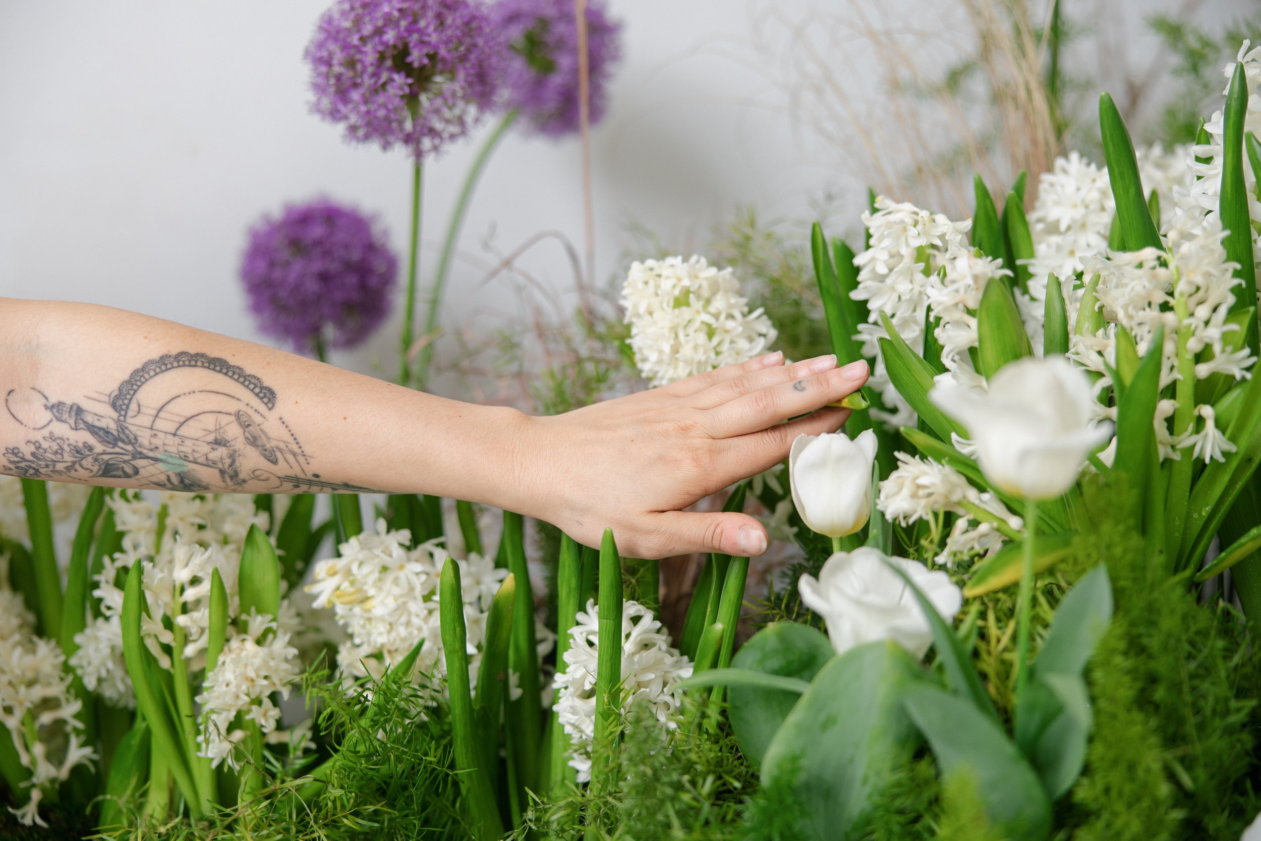 A person's arm with a tattoo resting among white and purple flowers in a garden.
