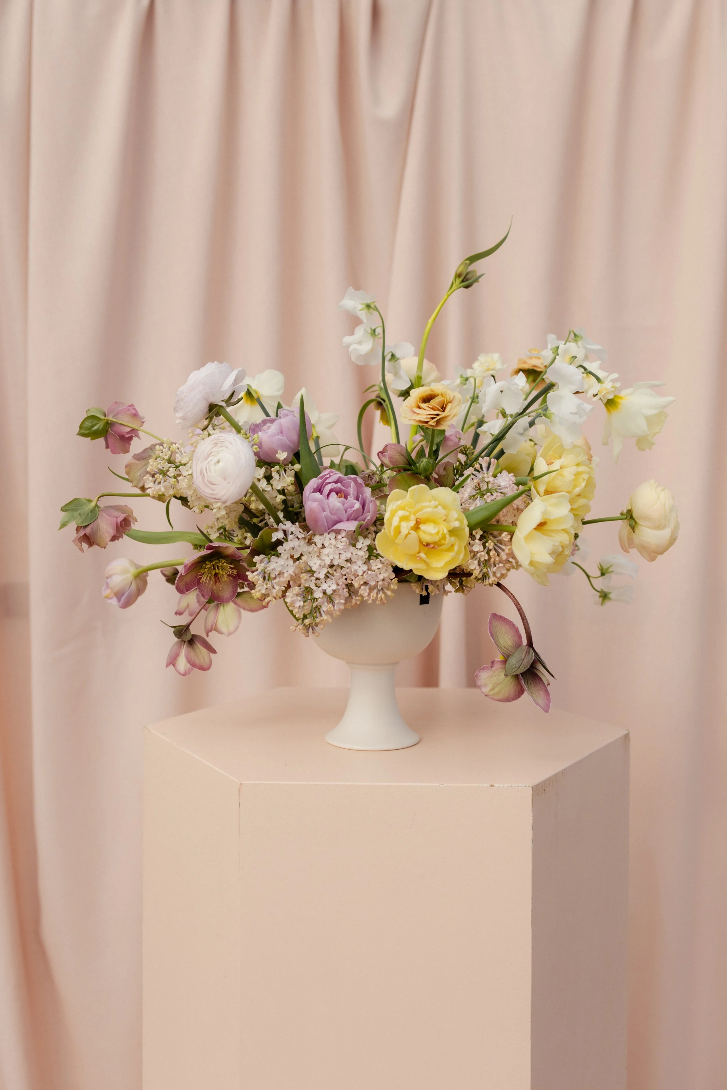 A floral arrangement with various white, pink, purple, and yellow flowers in a white pedestal vase against a light pink curtain background at The Minerva in Bath, UK with Trille Floral and Gaia Vessels.