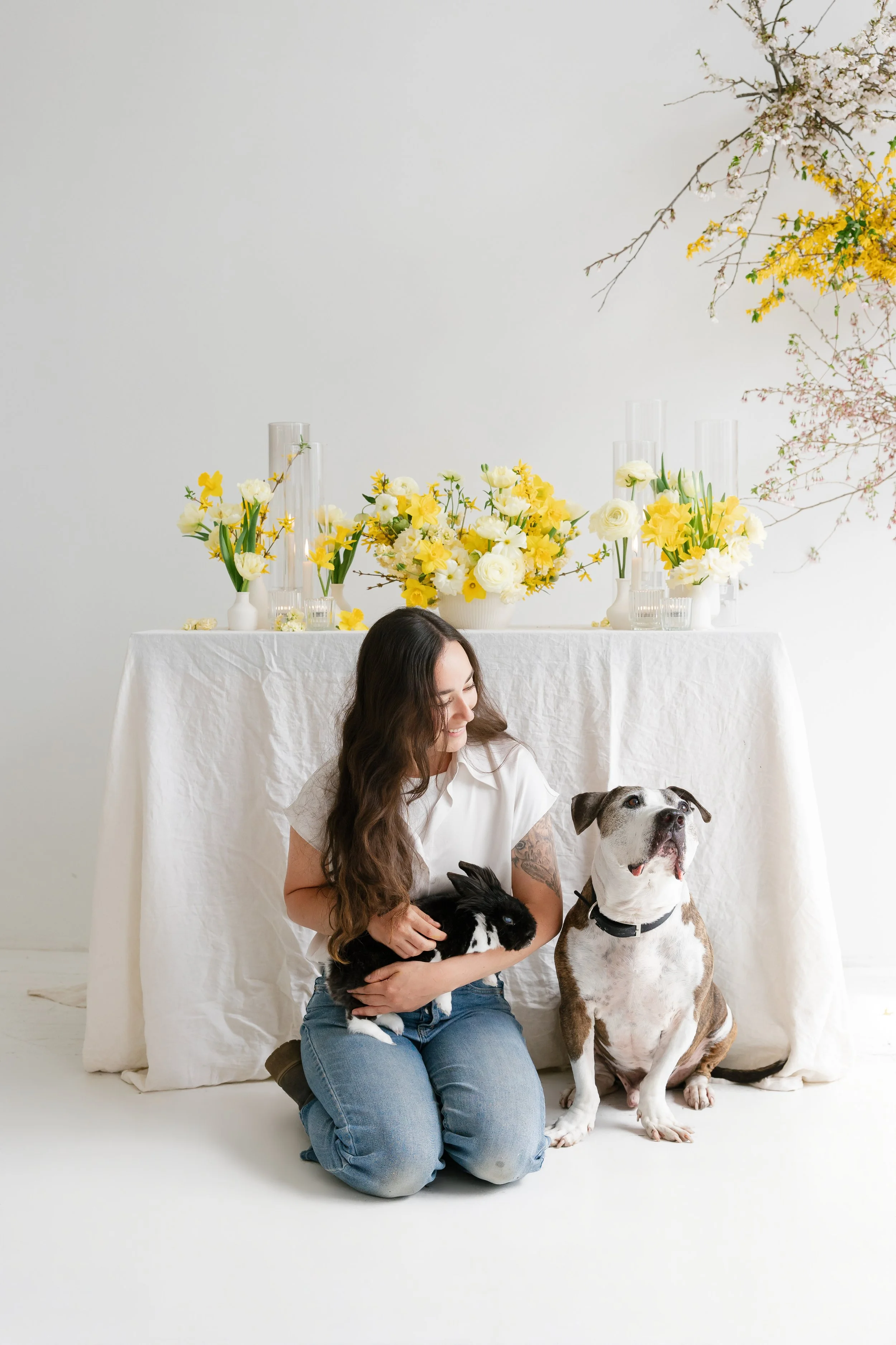 A woman with long brown hair sitting on the floor holding a black and white rabbit, with a large dog sitting beside her. They are in a white room with a background of a white table covered with a white cloth, decorated with yellow and white flowers a