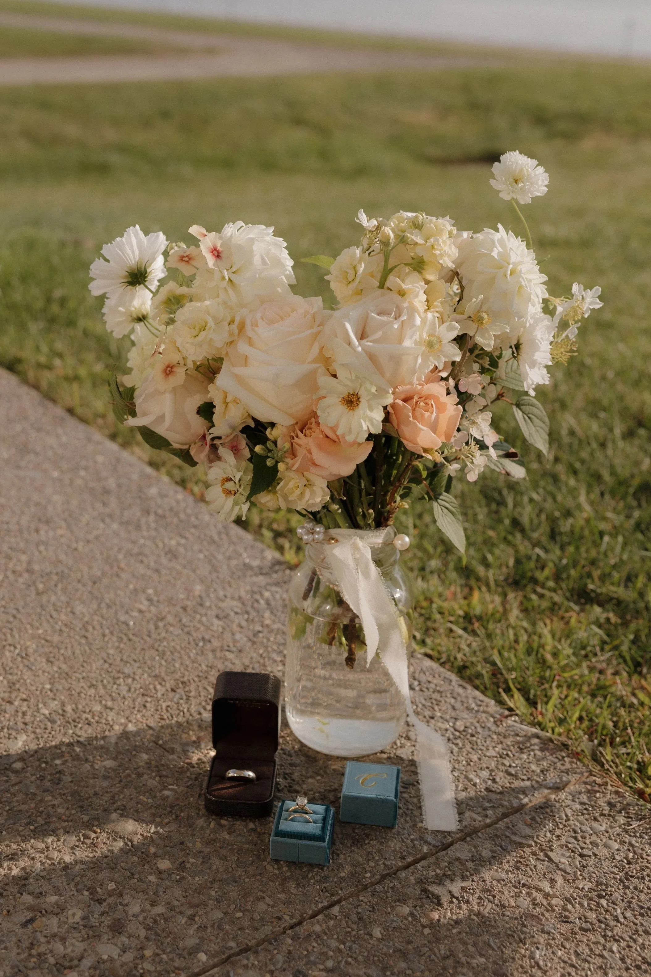 A bouquet of white and light-colored flowers in a glass vase, placed on a concrete surface outdoors, with jewelry boxes, a black ring box, and a small blue box with rings displayed. The background shows grass and a sidewalk near The Lake House.