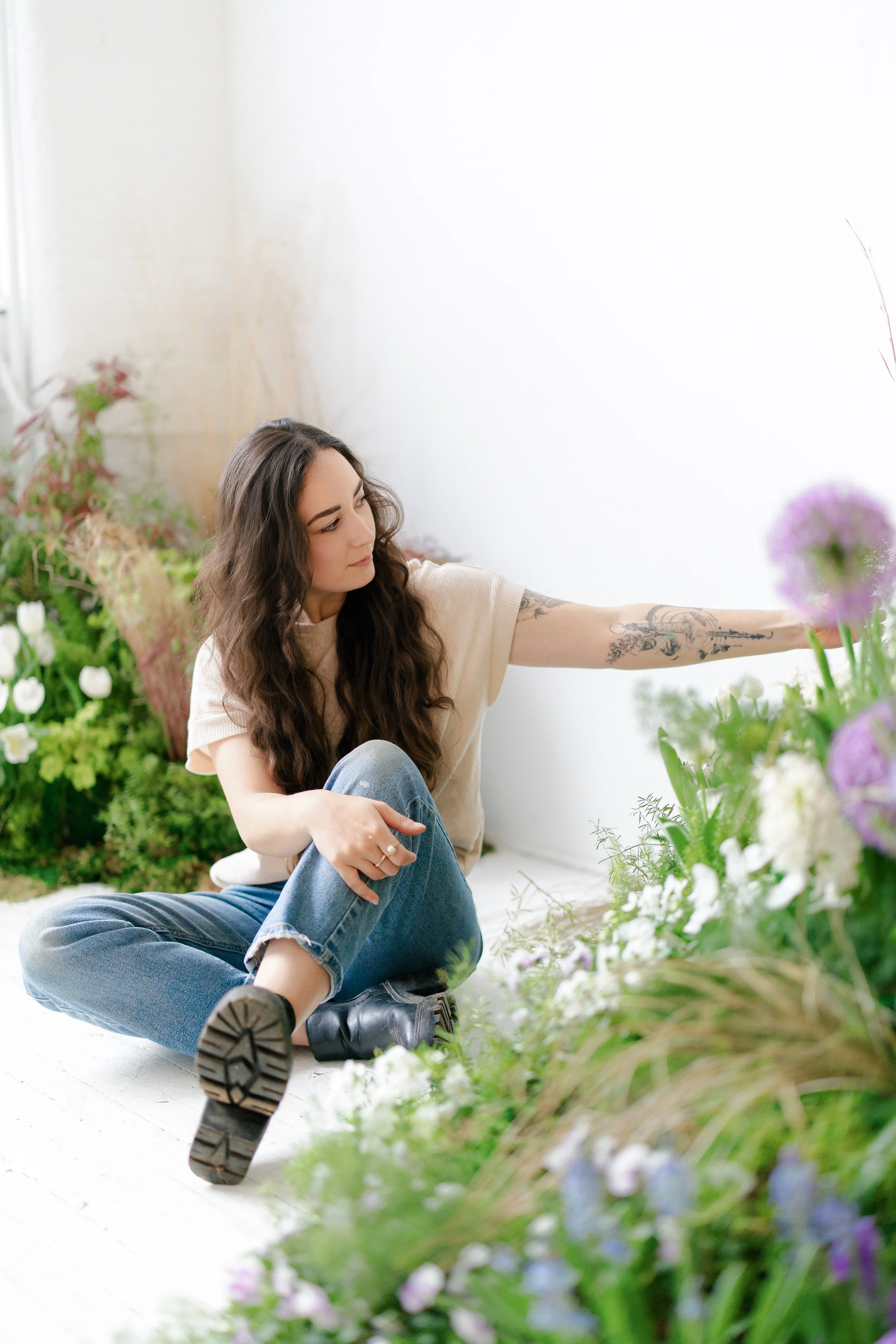 A woman with long, wavy brown hair and tattoos on her right arm sitting on the floor in a white room, reaching out to a flower arrangement of various white, purple, and green flowers and plants.