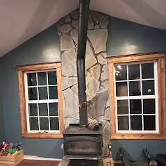 Cozy room with wood-burning stove and stone chimney between two windows.