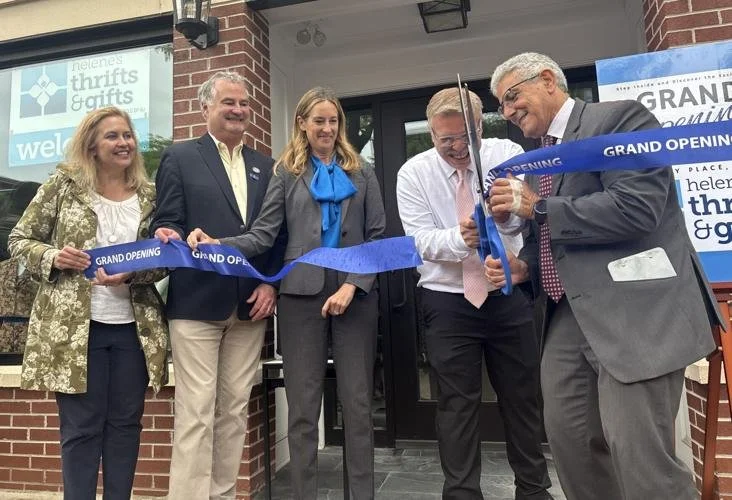Five people standing in front of a building, holding a blue ribbon that says 'Grand Opening'. One person is cutting the ribbon with scissors. They are celebrating the grand opening of a store or business.