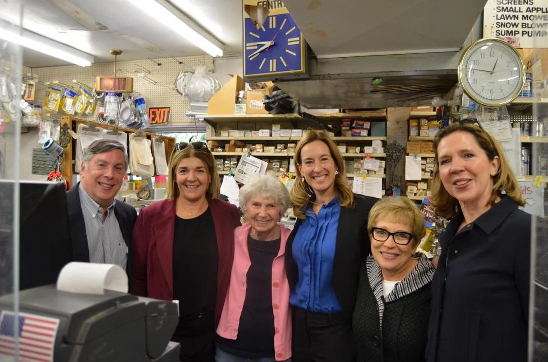 Six smiling people standing inside a store, with shelves and clocks visible in the background.
