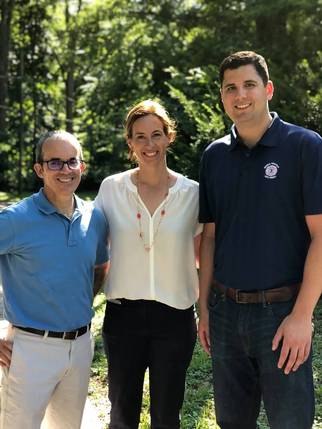 Three people standing outdoors on a sunny day, smiling, with trees and greenery in the background.