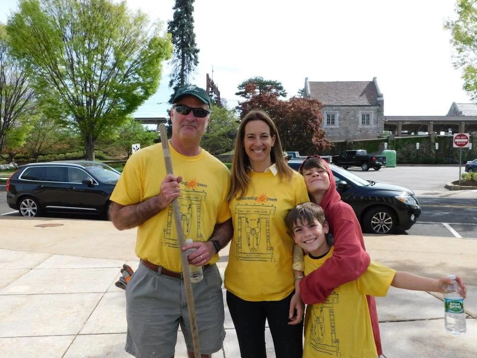 A group of four people standing outdoors on a sidewalk, wearing matching yellow T-shirts, with trees and buildings in the background.