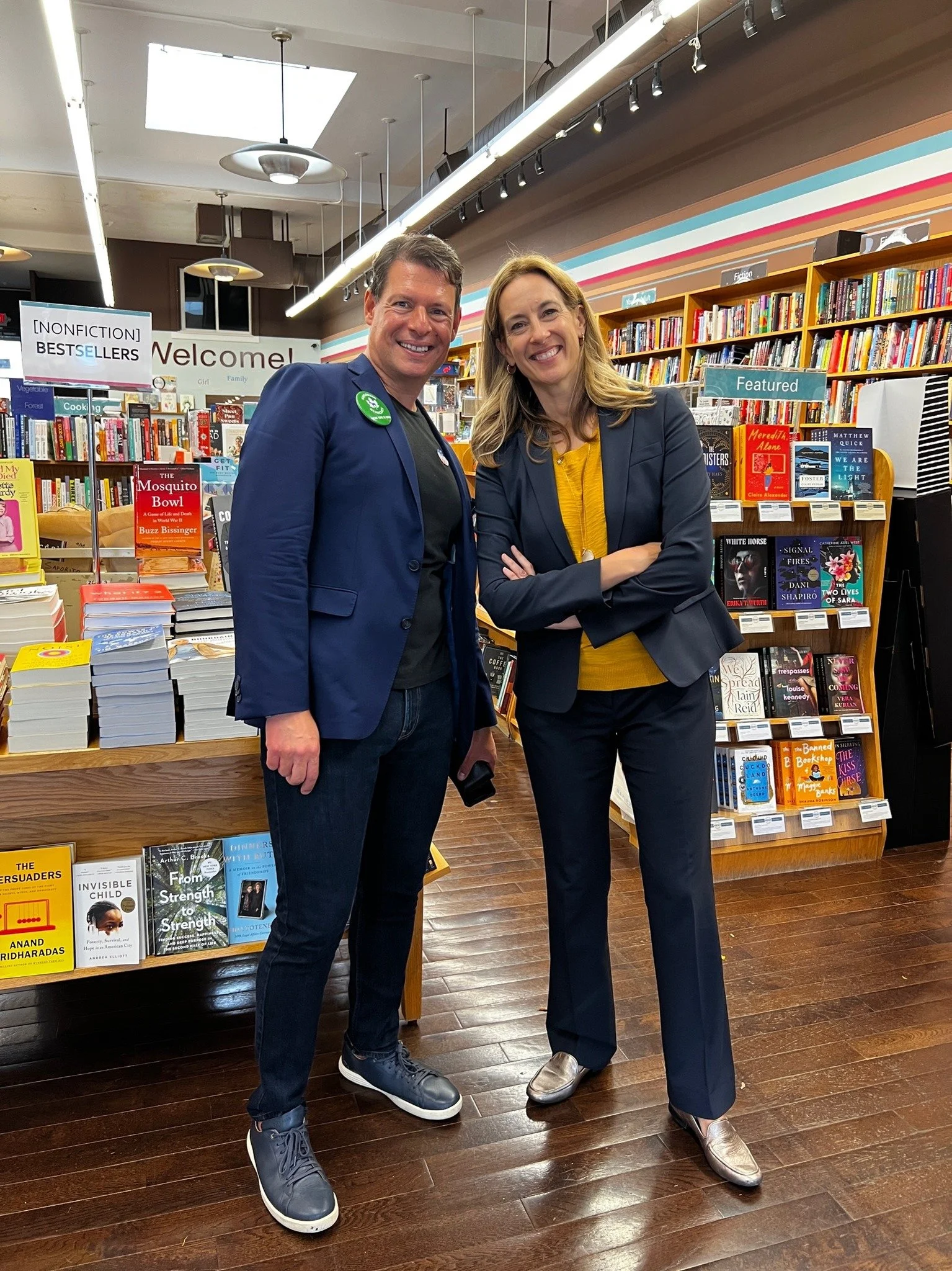 A man and a woman standing together inside a bookstore. The man is wearing a blue blazer, black shirt, and dark jeans. The woman is in a dark blazer, yellow top, and dark pants. Both are smiling, with the woman crossing her arms. Bookshelves filled w