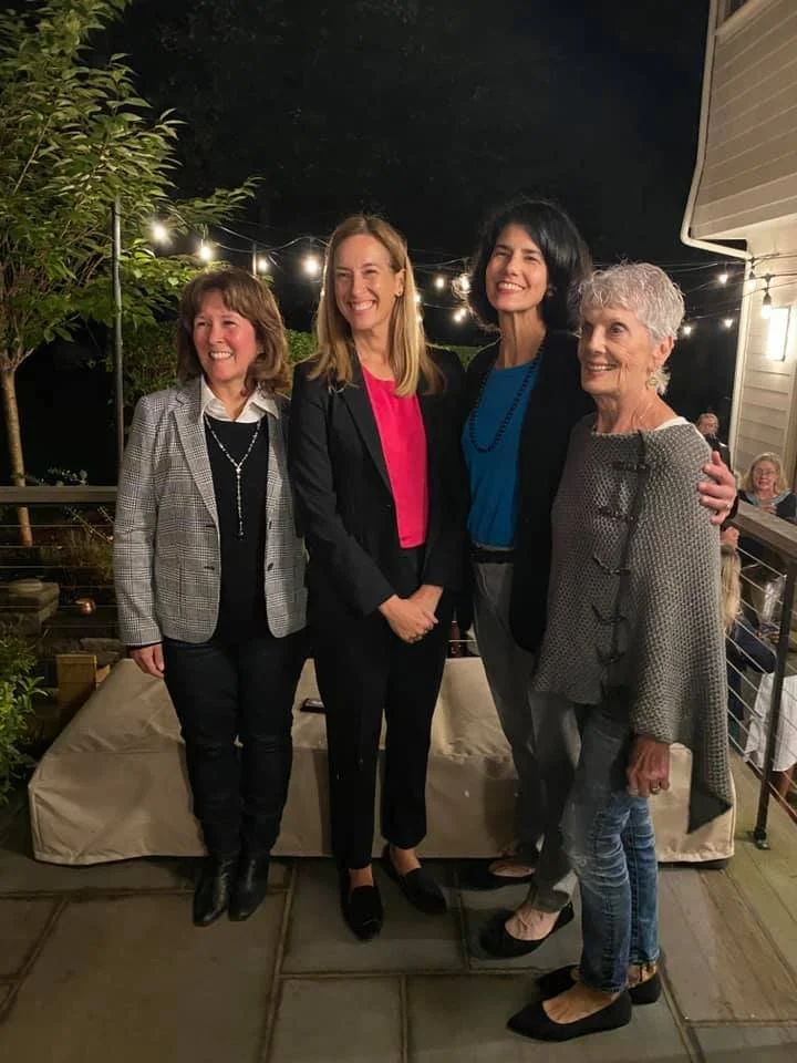 Four women standing together outdoors at night, smiling, with string lights overhead and a house wall to the right.
