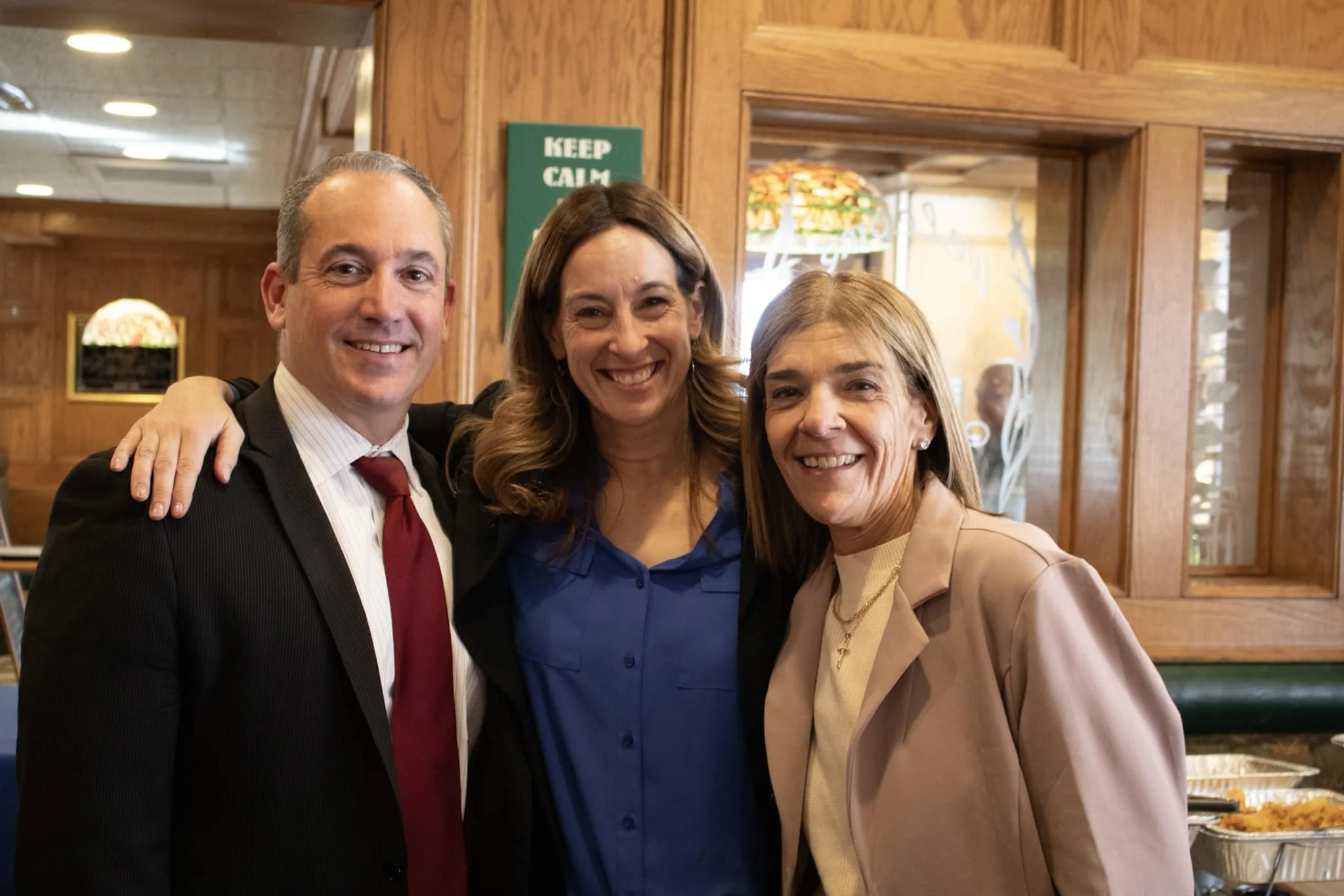 Three smiling adults, two women and one man, standing together with arms around each other in a restaurant.
