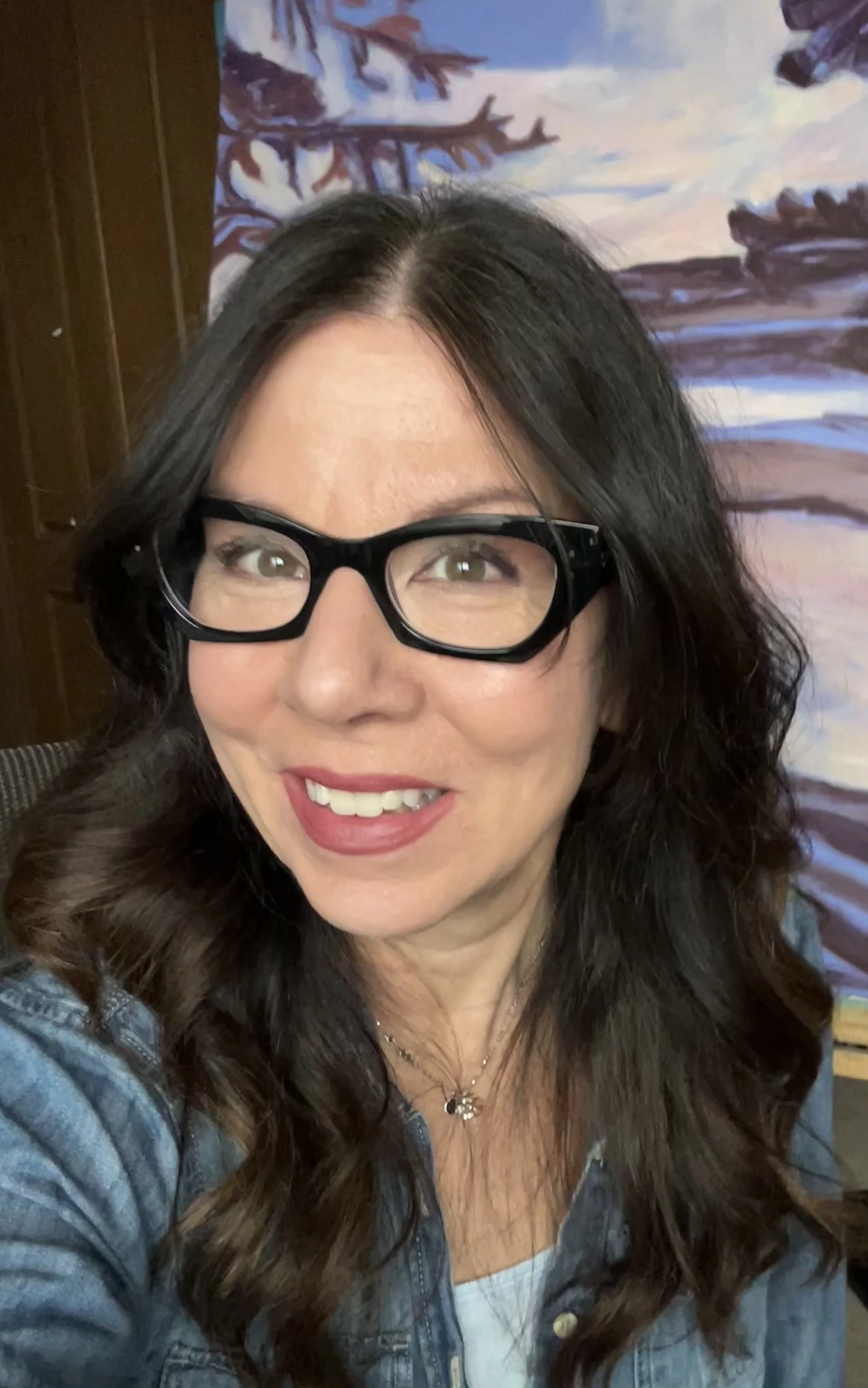 A woman with dark wavy hair, wearing glasses and a denim jacket, smiling for a photo with a colorful landscape painting in the background.