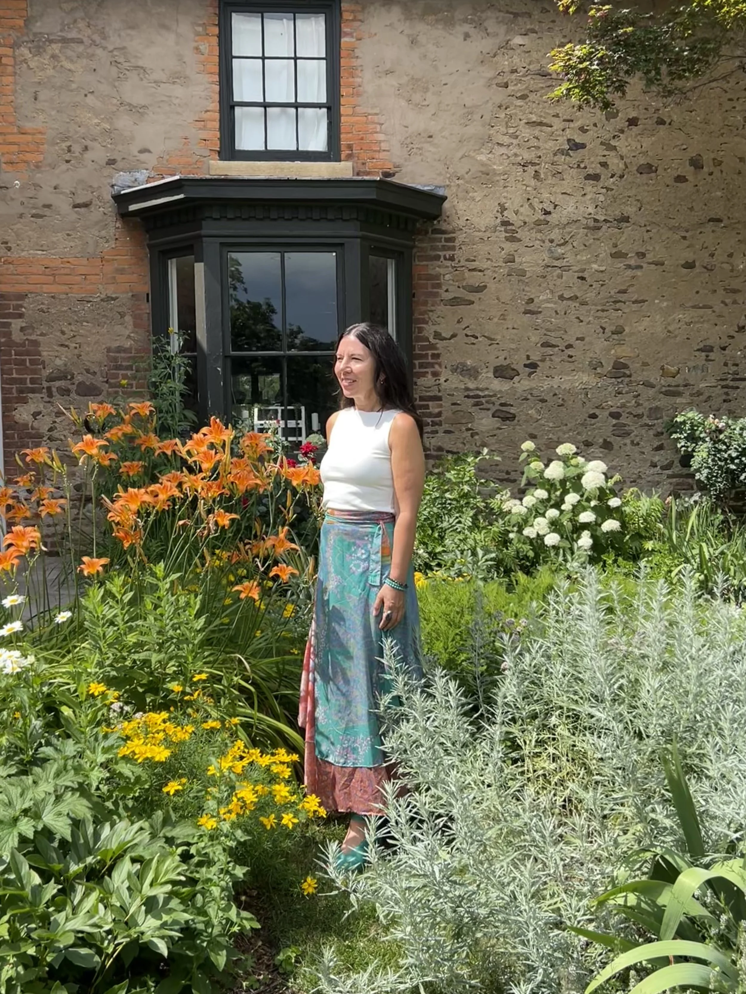 A woman standing in a garden with colorful flowers and greenery, in front of a brick building with a large window and black trim.