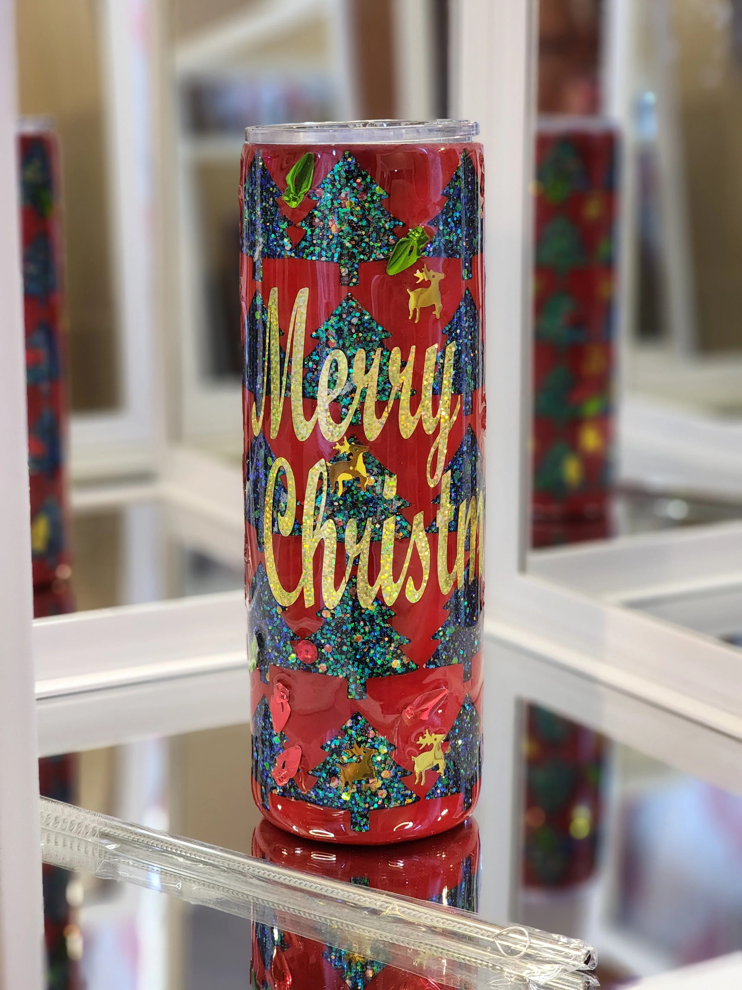 A red Christmas-themed tumbler decorated with glittery blue trees, gold reindeer, and the words "Merry Christmas" in gold cursive.