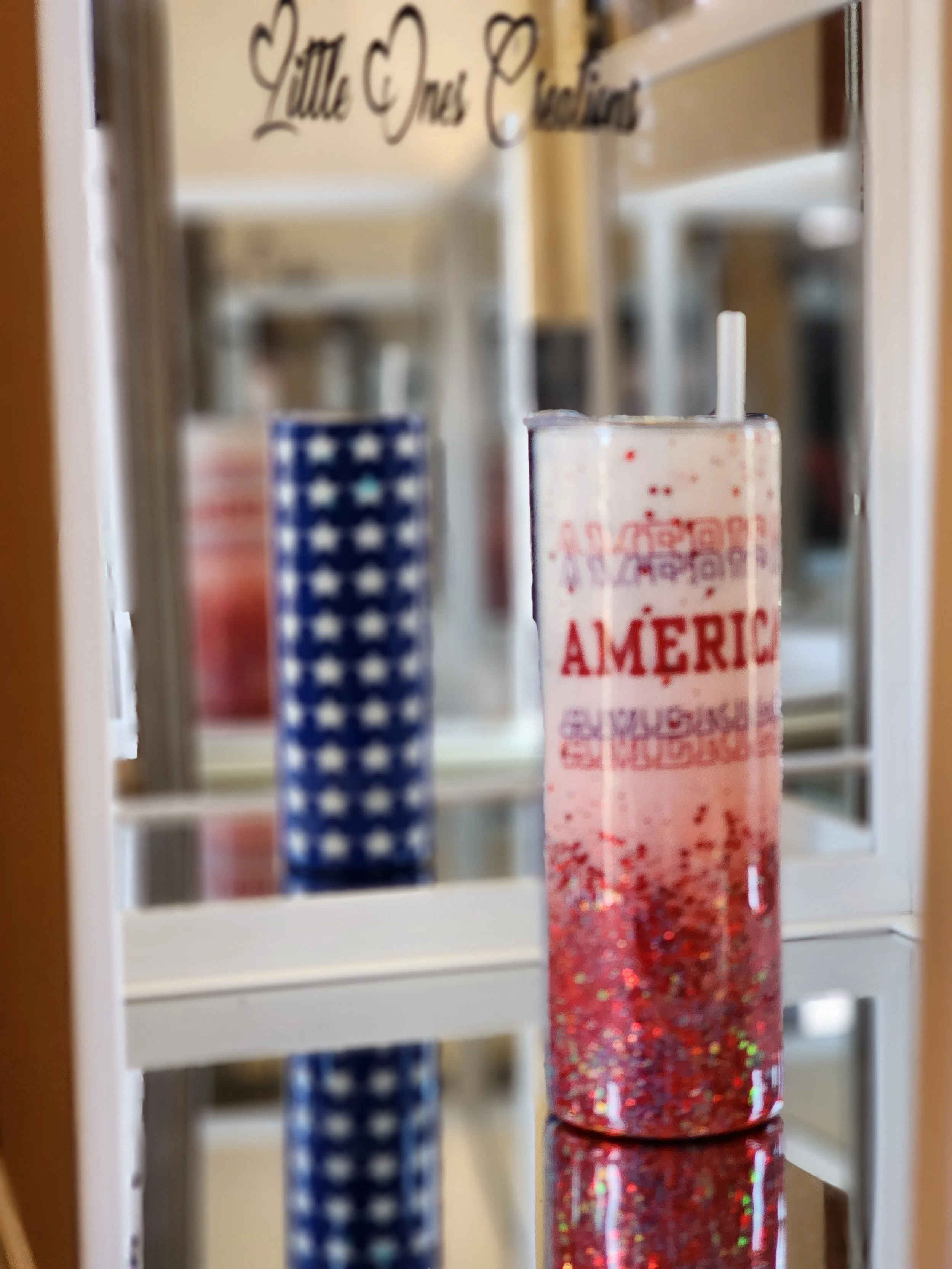 A glass of pink cocktail with a straw, decorated with red, white, and blue glitter, placed on a mirrored surface, with patriotic decorations and a sign reading 'Little One Clubhouse' in the background.