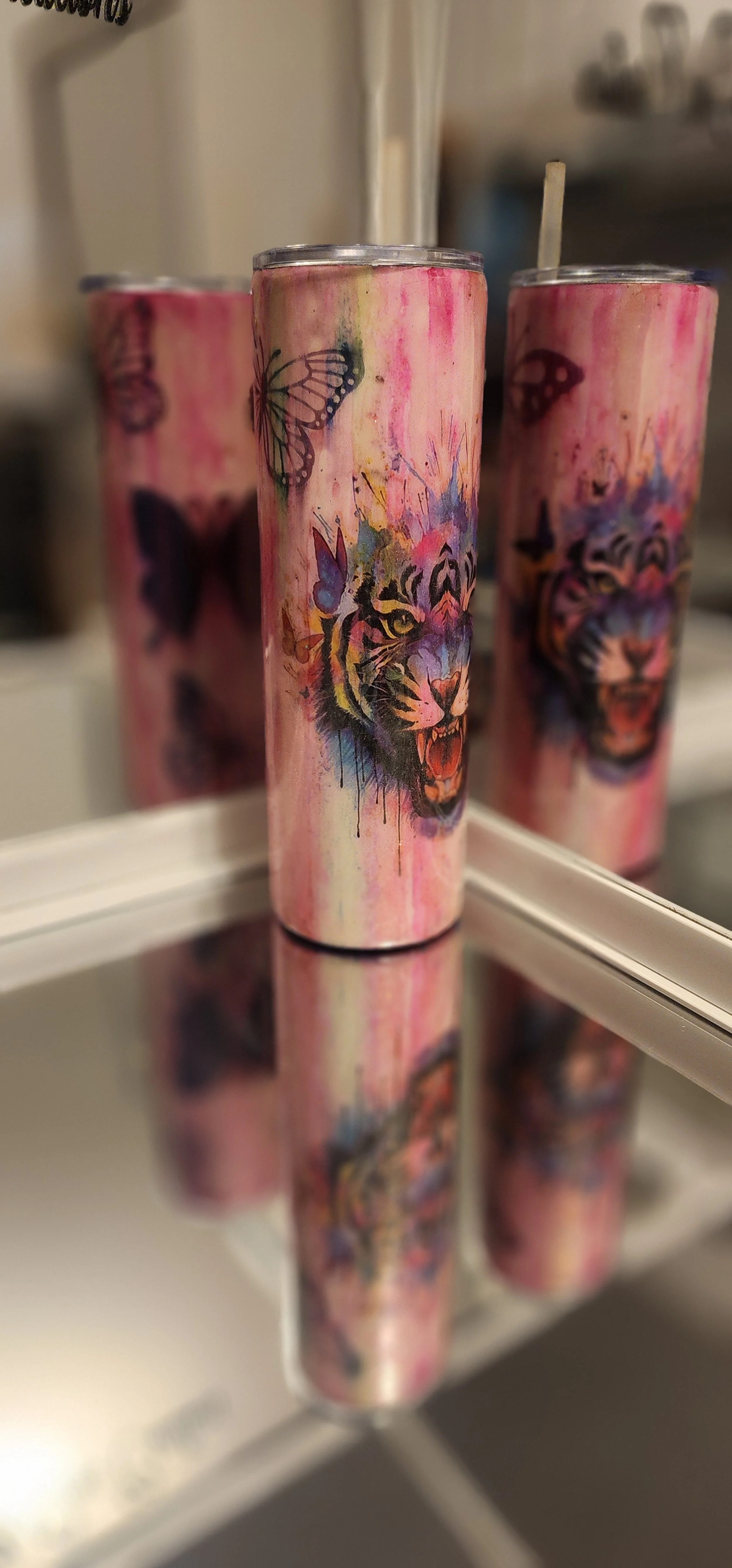 Three tall cans with colorful animal designs, including a tiger, butterflies, and abstract splashes, reflected on a mirror surface.