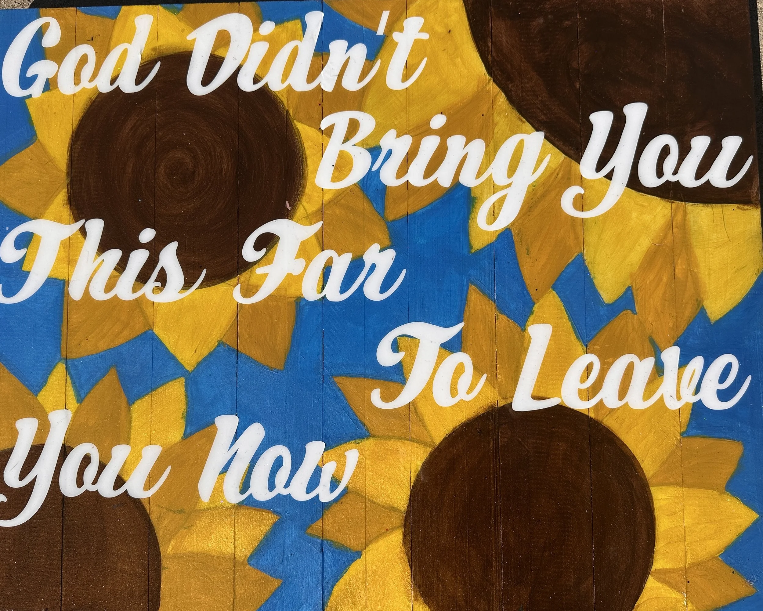 A colorful painted wooden sign with sunflowers and a quote reading, "God Didn't Bring You This Far To Leave You Now."
