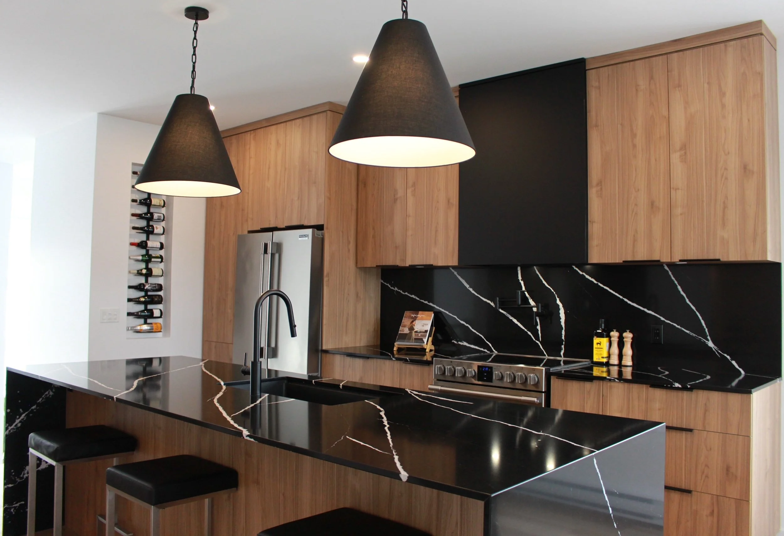 Cuisine moderne avec comptoir en marbre noir, armoires en bois clair, tabourets noirs, éclairage suspendu et réfrigérateur en acier inoxydable.