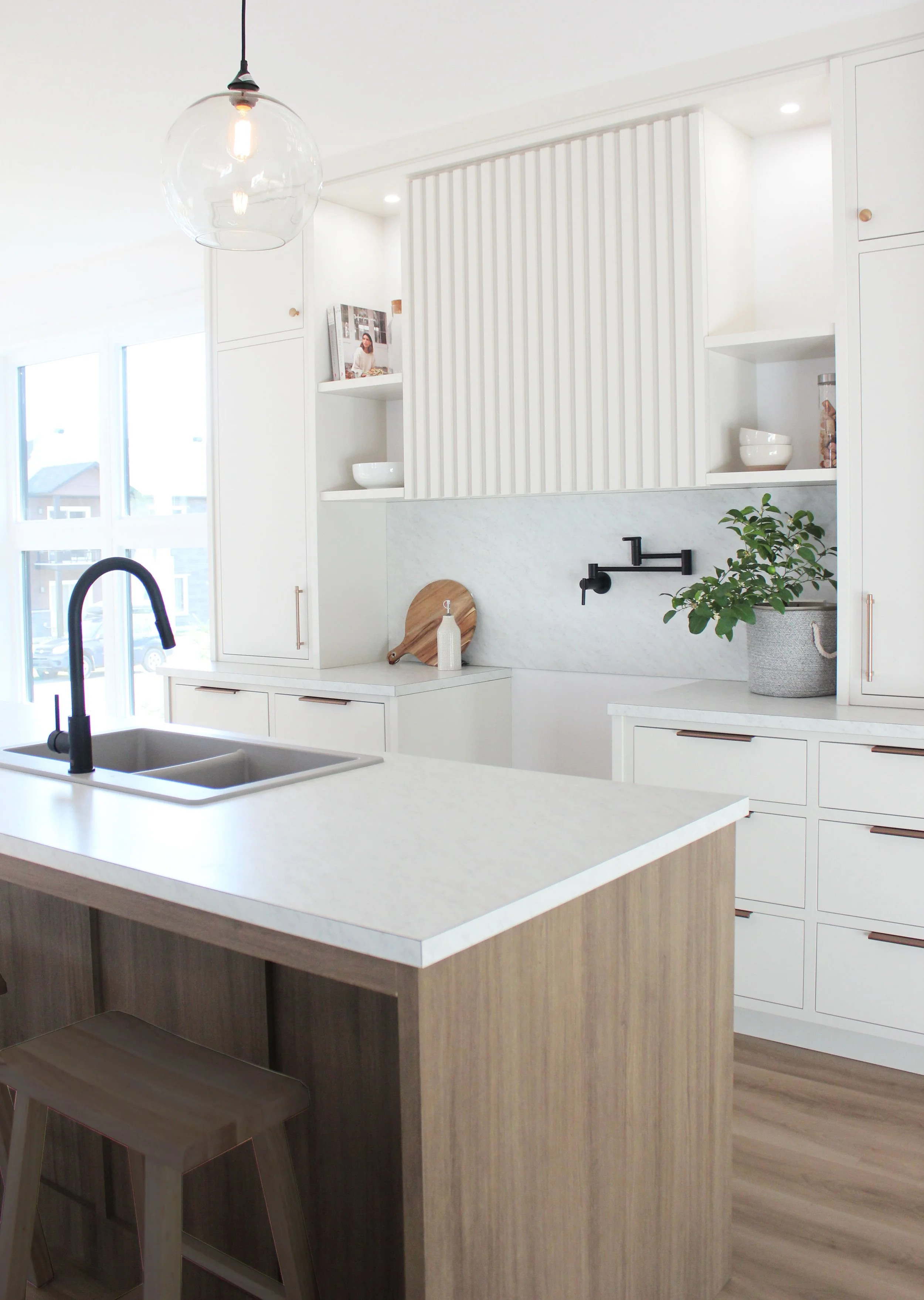 Cuisine moderne avec un îlot central en bois, des armoires blanches, un évier double avec robinet noir, une plante en pot, des planches à découper et un luminaire suspendu transparent.