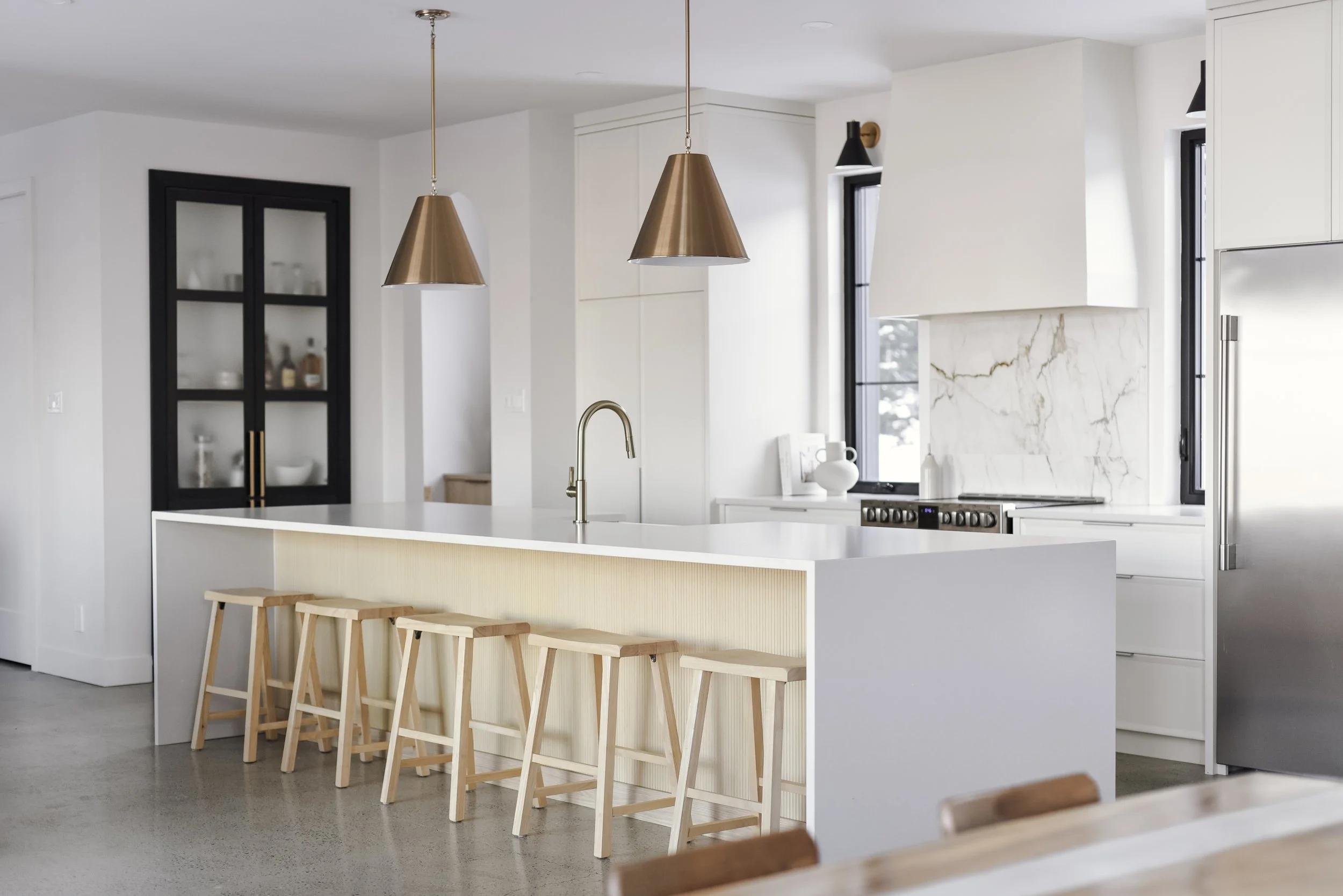 Cuisine moderne avec îlot central, tabourets en bois, luminaires suspendus en métal, et décoration minimaliste.