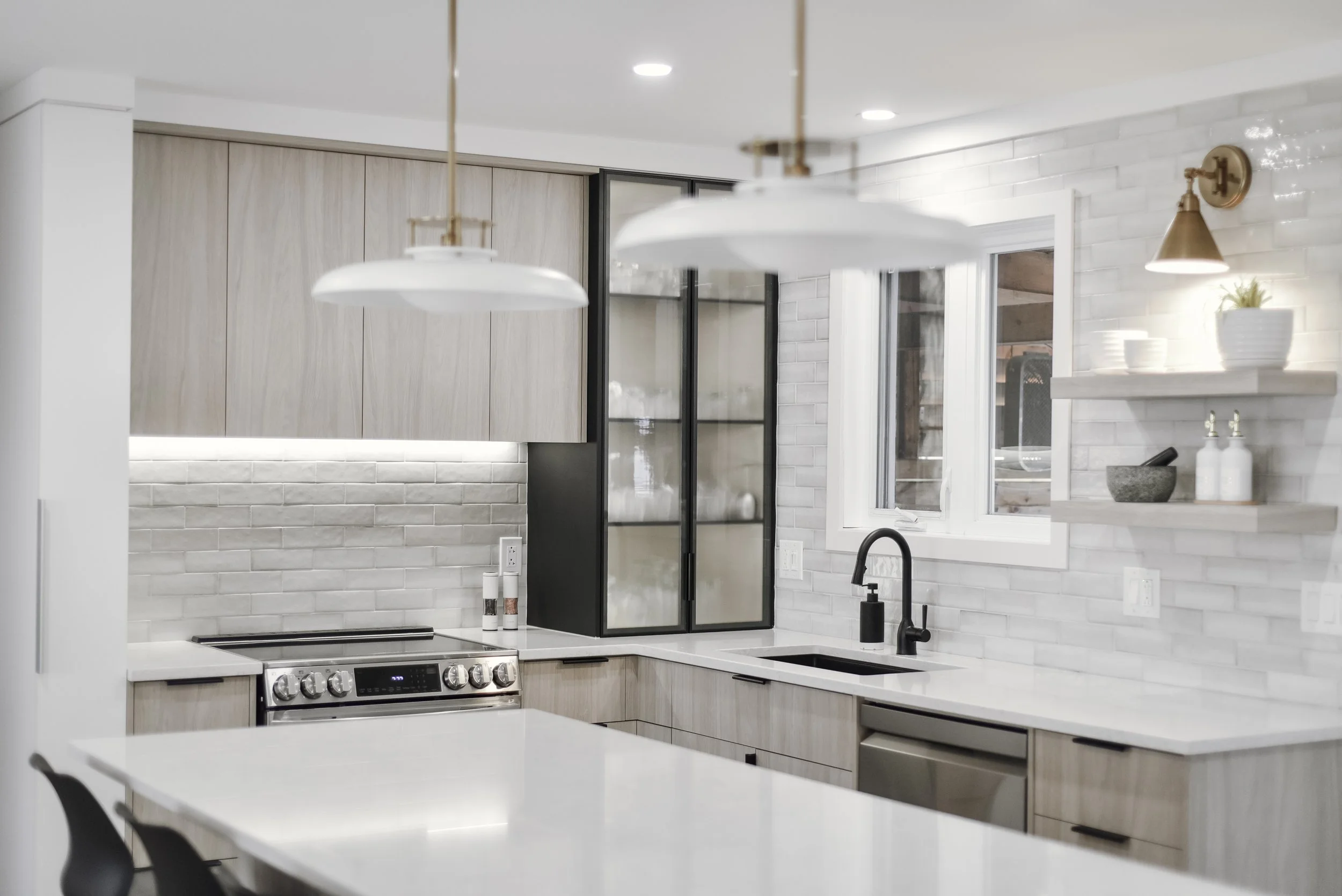 Cuisine moderne avec îlot, armoires en bois clair, plan de travail en marbre blanc, cuisinière en acier inoxydable, évier noir, étagères ouvertes avec vaisselle.