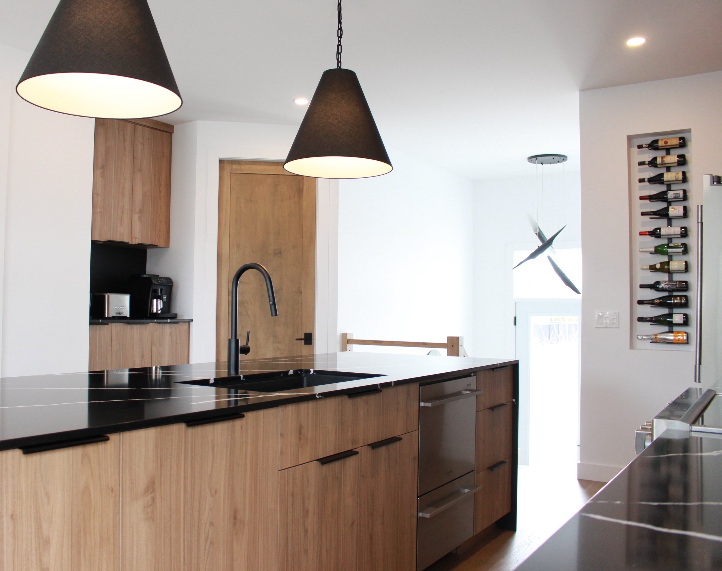 Cuisine moderne avec îlot central et comptoirs en bois clair, évier noir et électroménagers en acier inoxydable. Deux lampes suspendues au-dessus de l’îlot. Armoire murale avec bouteilles de vin.