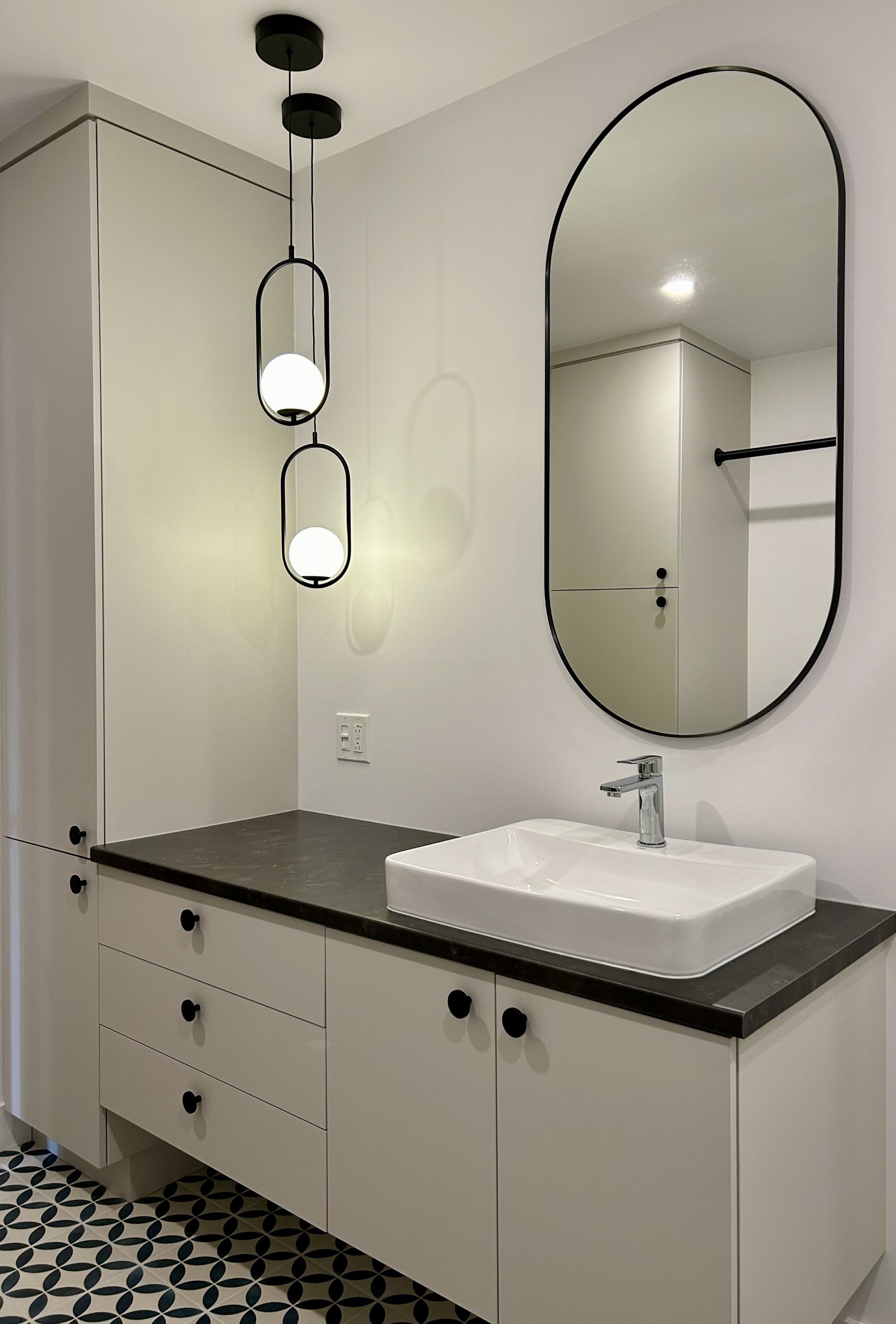Salle de bain moderne avec un évier rectangulaire sur un comptoir sombre, un grand miroir ovale, des lampes suspendues design et des placards blancs.