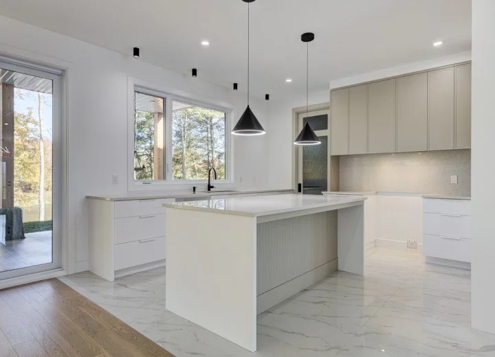 Cuisine moderne avec îlot central, éclairage suspendu, sol en marbre et armoires blanches. Grande fenêtre avec vue sur l'extérieur.