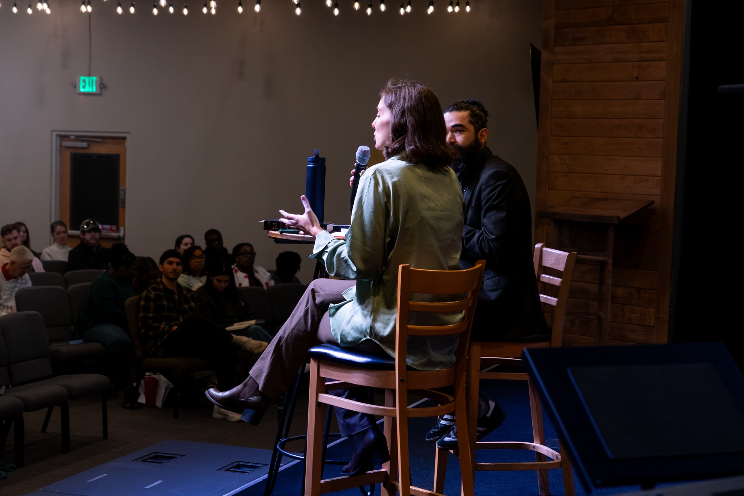 A woman speaking into a microphone while sitting on a stage in front of an audience, with a man sitting beside her. The audience is seated in a darkened room, and the background features wooden paneling on the wall.