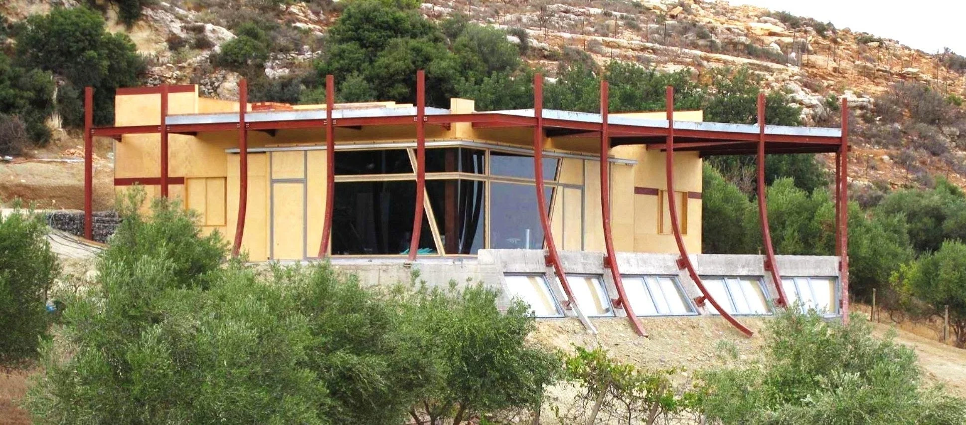 Modern house under construction on hillside with curved red beams, large windows, surrounded by green trees and shrubbery.