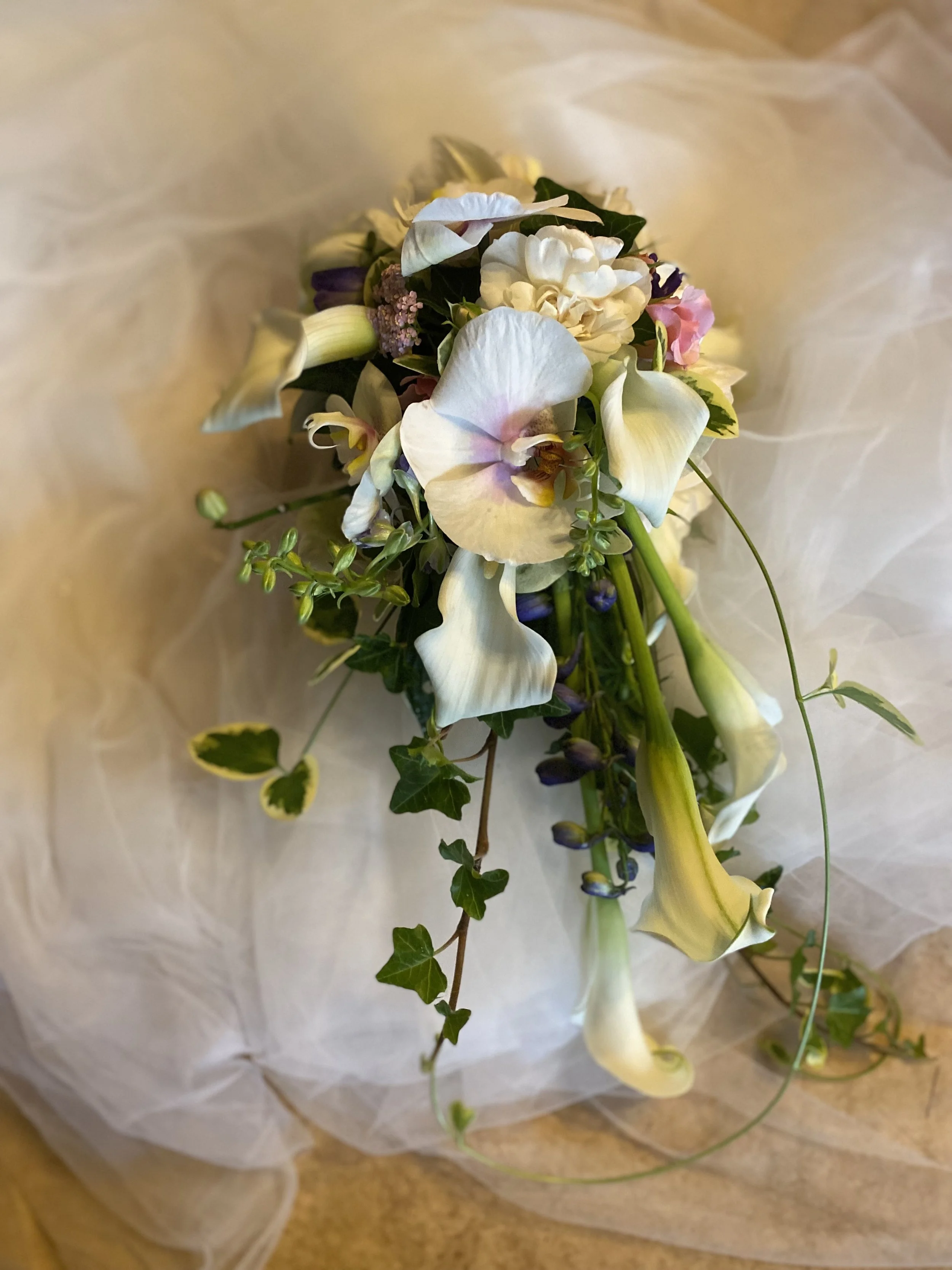 Bouquet de fleurs blanches, dont des orchidées, avec des feuilles vertes, posé sur un tissu blanc et un sol en bois.