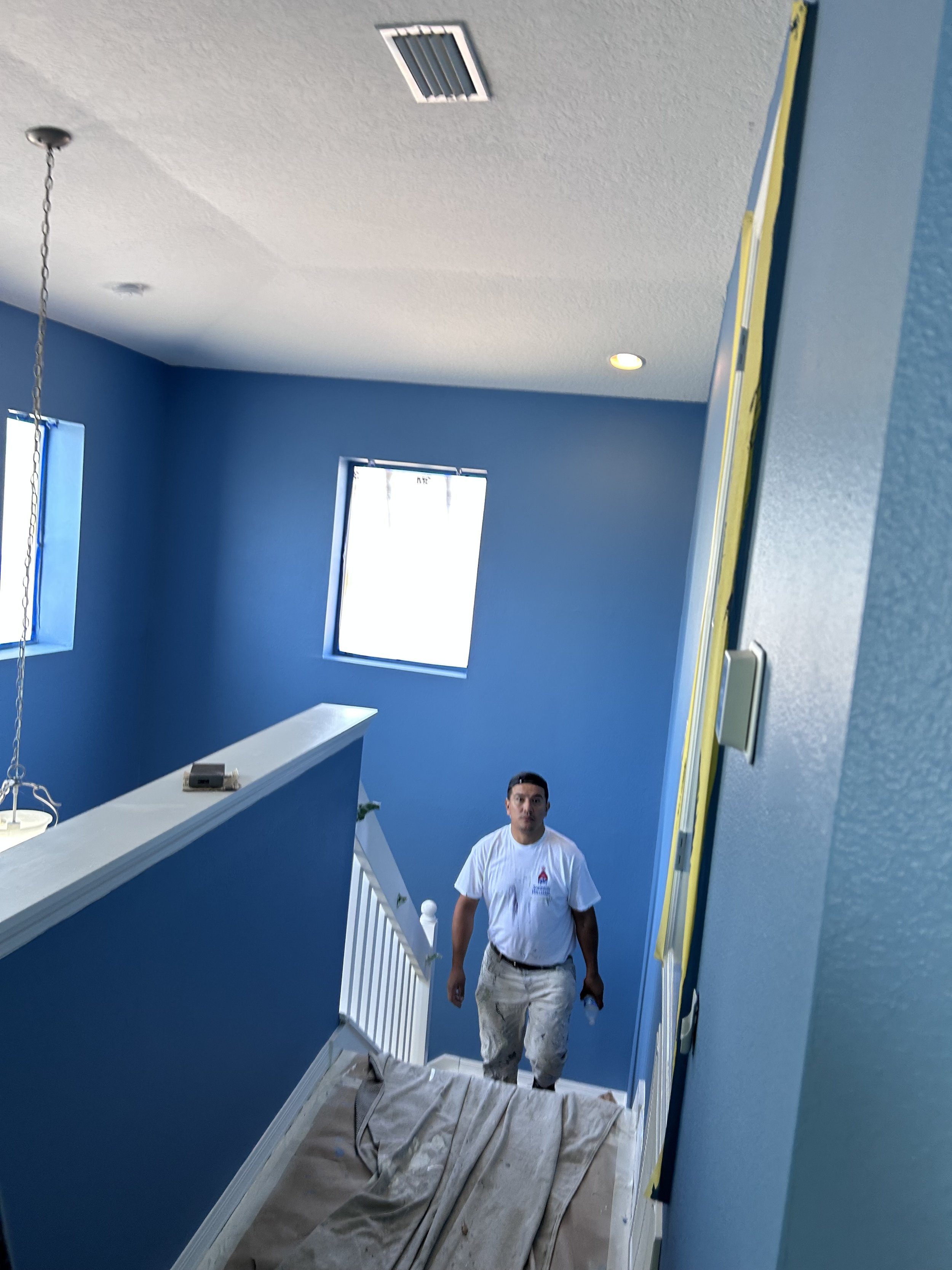 A painter in a white shirt and paint-splattered pants standing on a stairway covered with a protective tarp, in a room with blue walls and two windows.