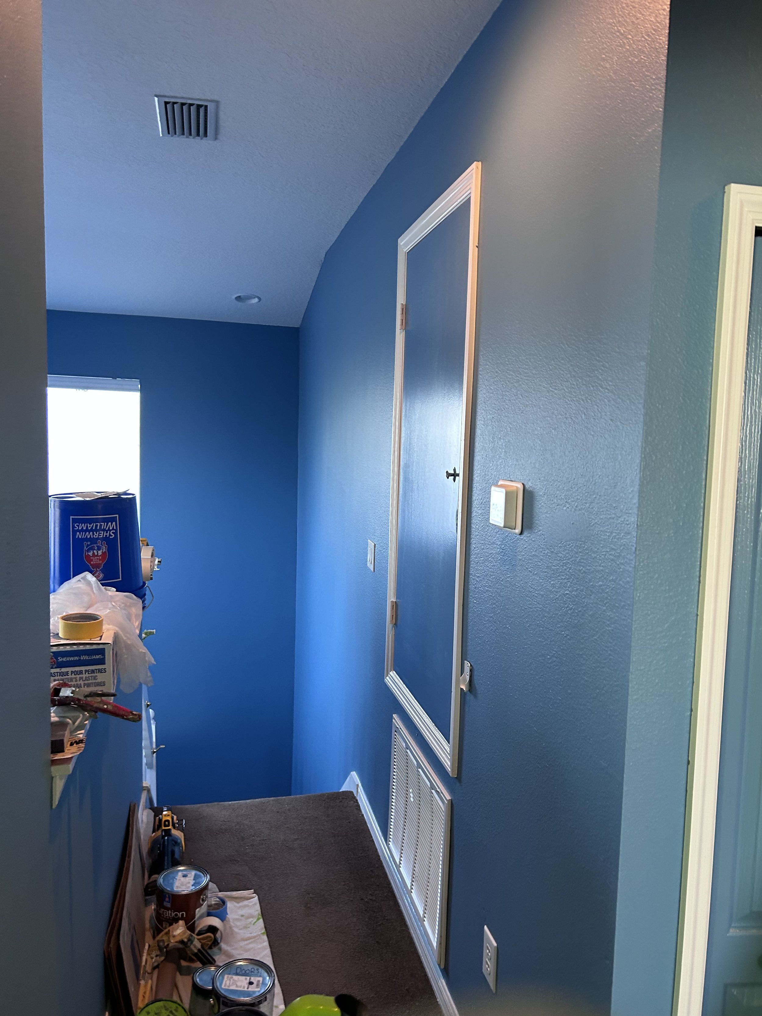 Interior hallway in a home with blue walls and a ventilation panel, painting supplies stacked on a shelf.