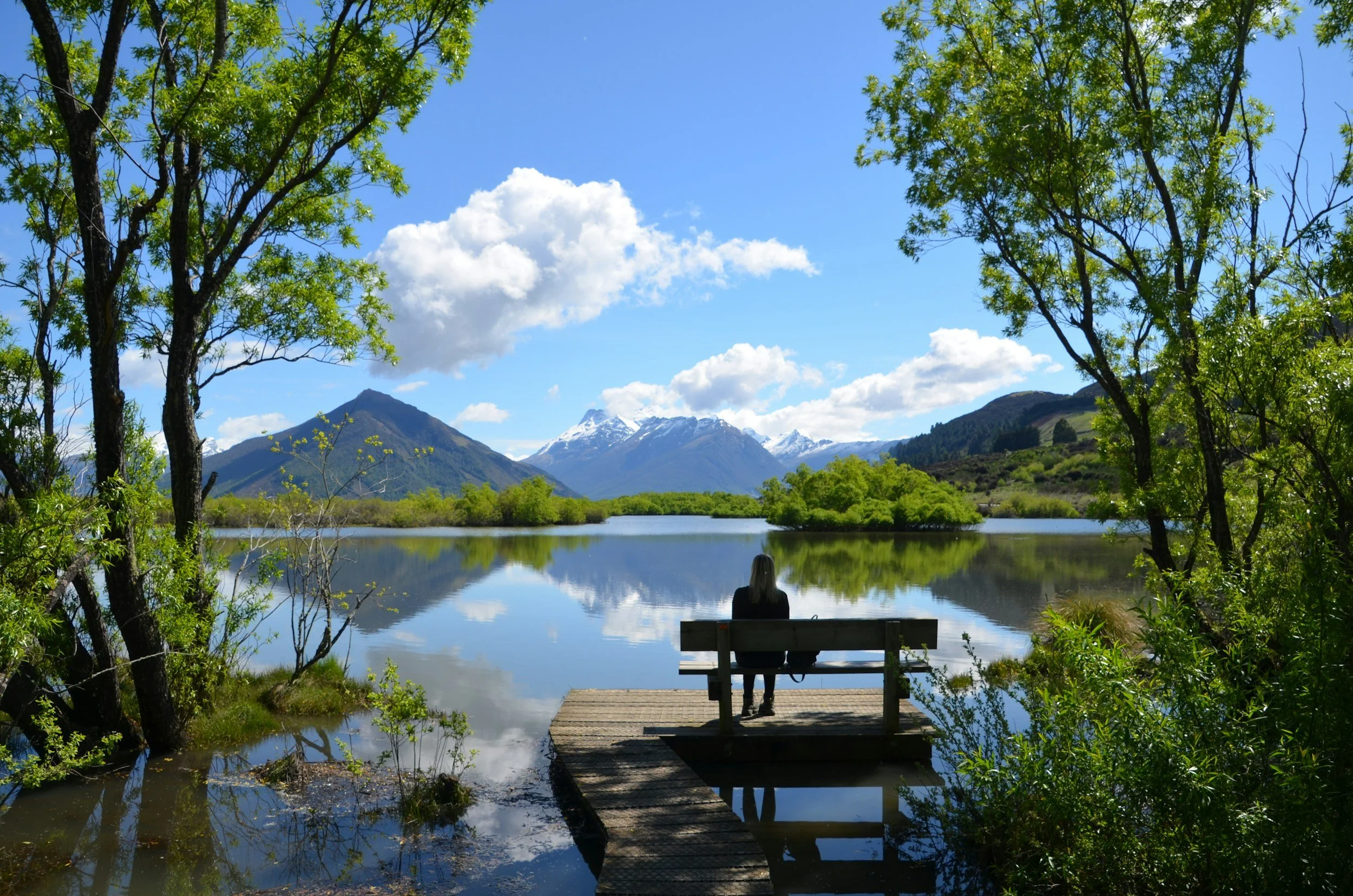 A woman sitting on a wooden dock by a calm lake with mountains and snow-capped peaks in the background, surrounded by green trees and blue sky with clouds.