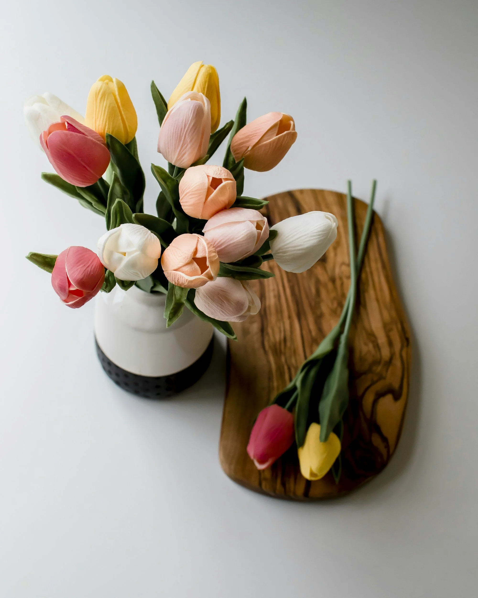 A bouquet of multicolored tulips in a white vase and one yellow and pink tulip on a wooden tray.